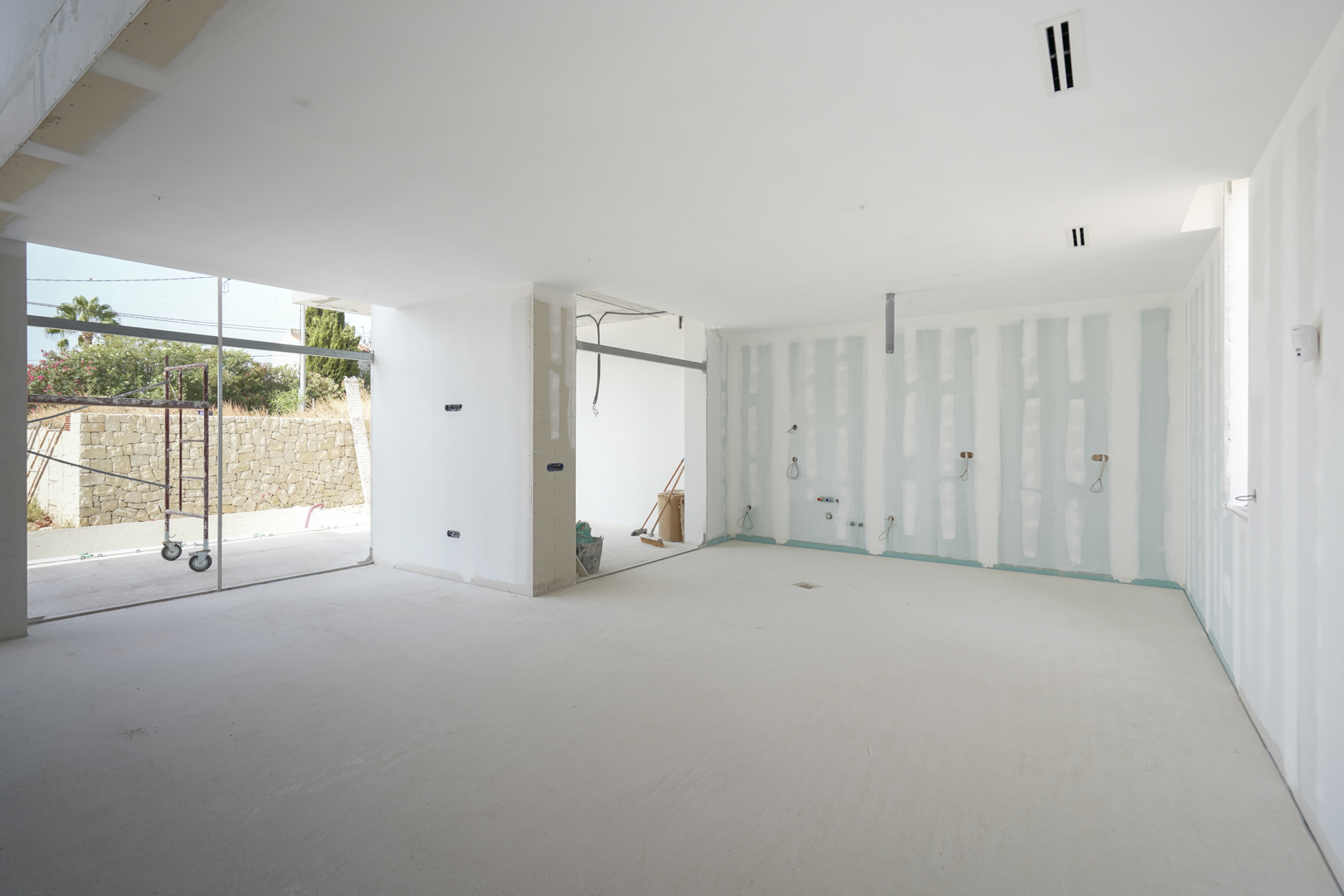Interieur van de woonkamer in aanbouw, voorzien van plafondroosters voor airconditioning en grote raampartijen die uitkijken op de tuin.