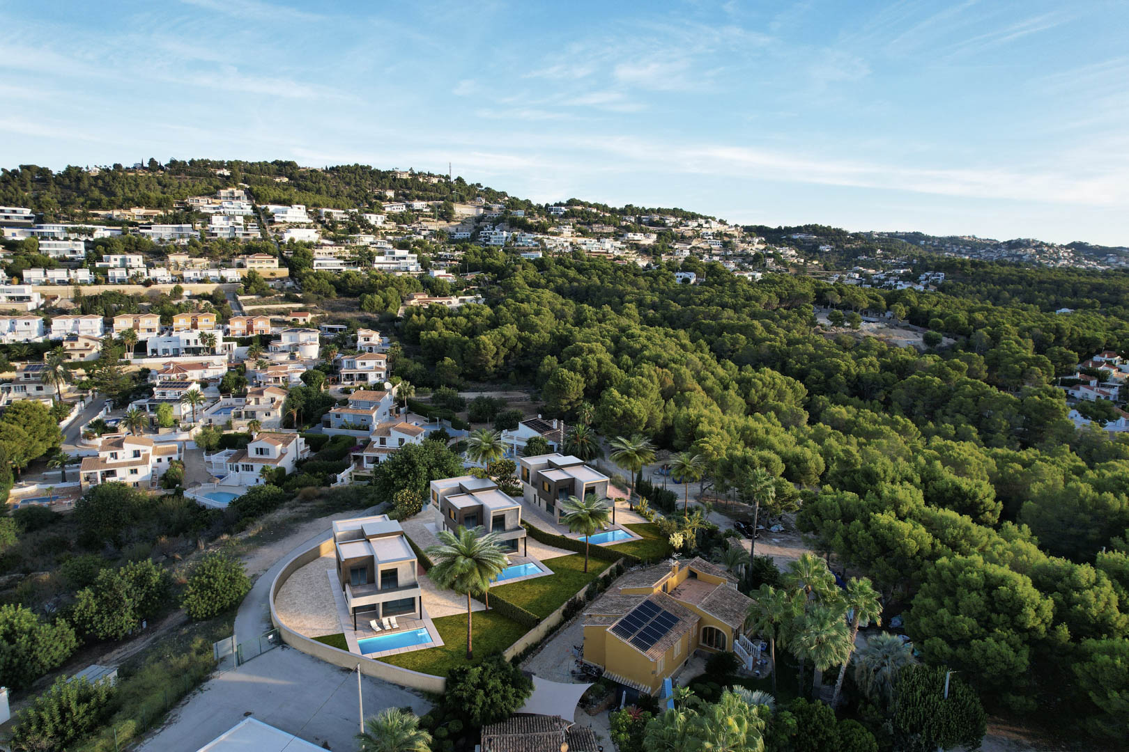Luchtfoto van de drie moderne villa's met privézwembaden en tuinen, gelegen op een heuvel grenzend aan een uitgestrekt dennenbos in Calpe.
