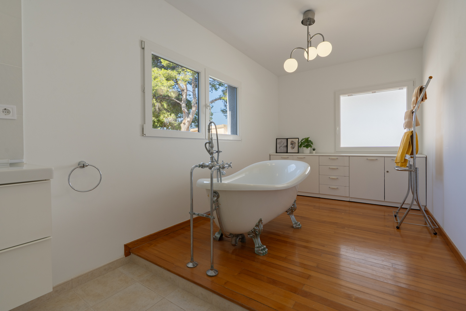 Badkamer met een vrijstaand bad op pootjes op een houten platform, voorzien van witte kasten en ramen met uitzicht op de mediterrane tuin.