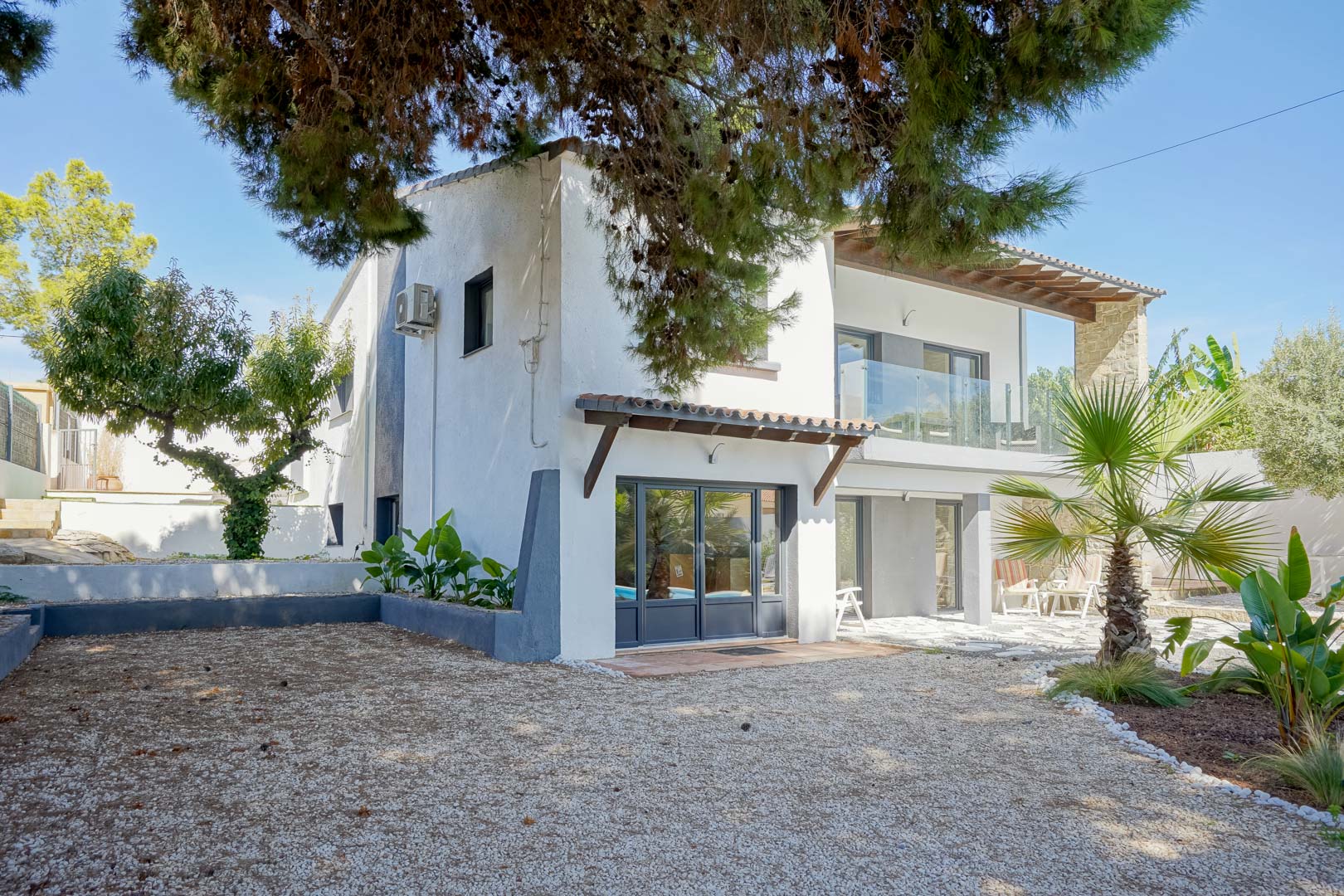 Buitengevel van de villa met grijze kozijnen, terrassen met glazen balustrades en een mediterrane tuin met grind en volwassen pijnbomen.