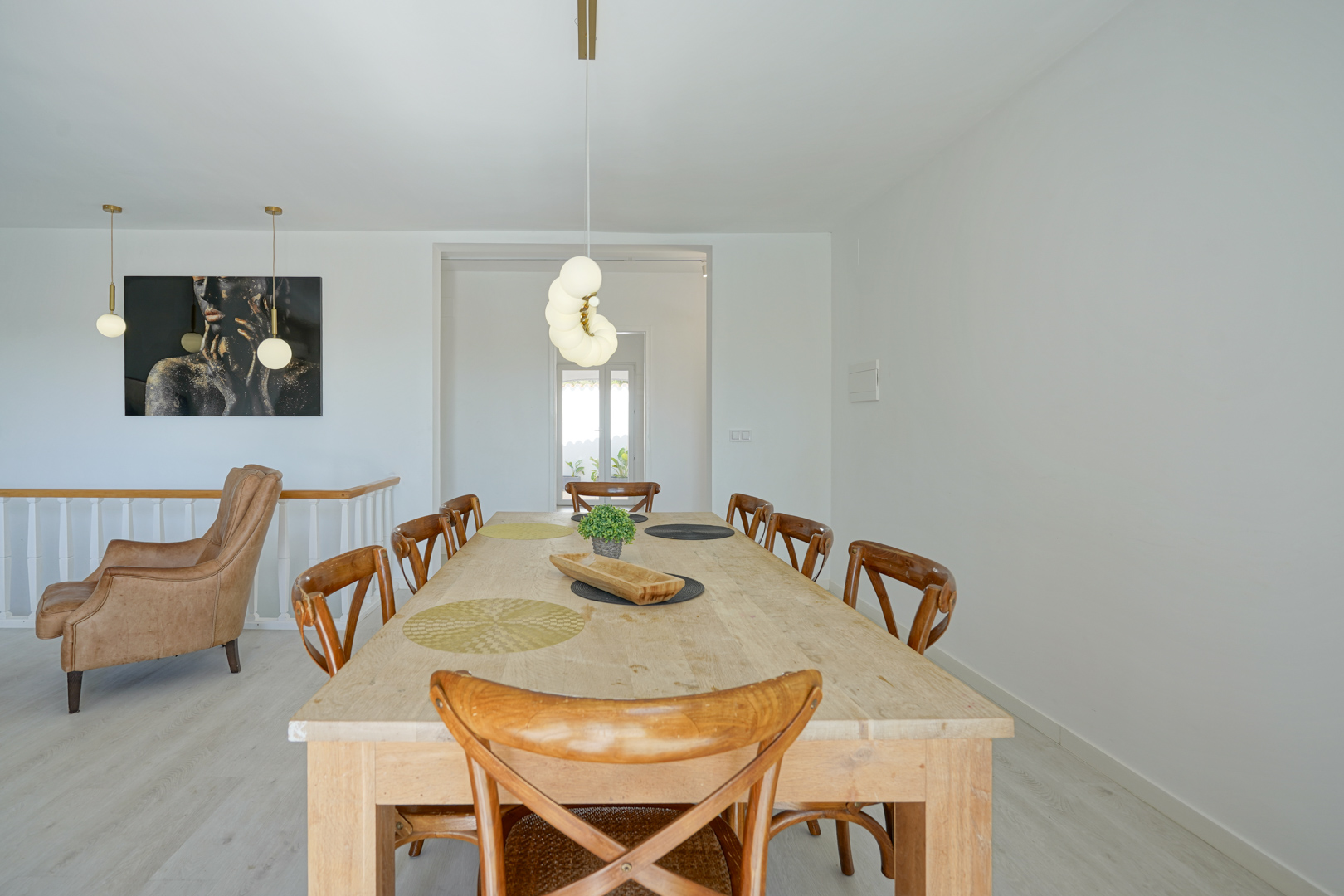 Eethoek met een grote licht houten tafel, kruisrugstoelen en moderne bolvormige hanglampen. Op de achtergrond is een leren fauteuil en een kunstzinnig schilderij zichtbaar.