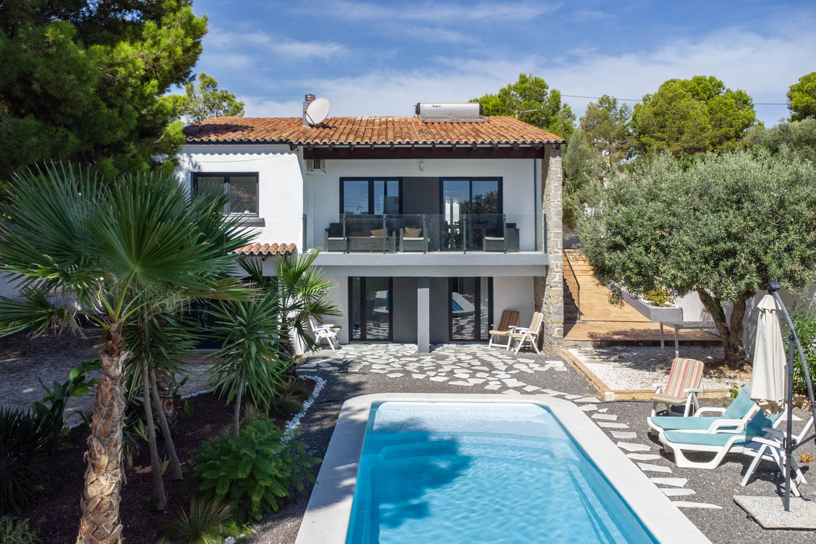 Exterieur van de villa met rechthoekig zwembad, mediterrane tuin en witte gevel. Voorzien van glazen terrasbalustrades en een zonneboiler op het pannendak.