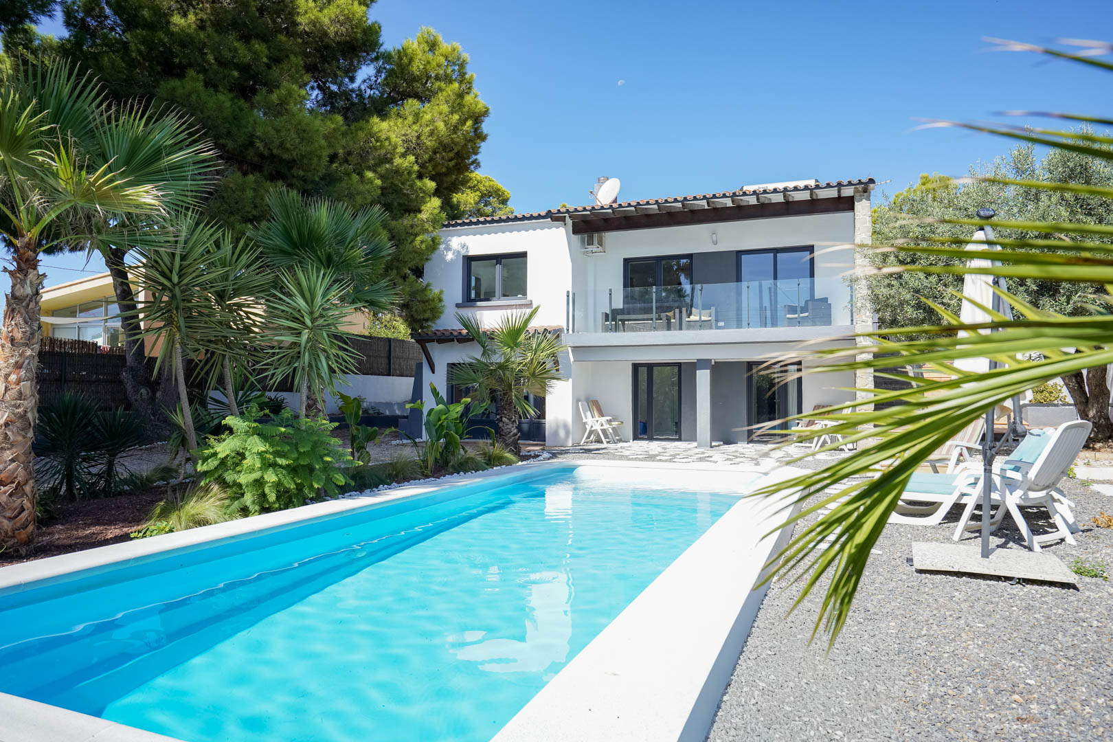 Exterieur van de villa met rechthoekig zwembad, mediterrane tuin met palmbomen en een witte gevel van twee verdiepingen met glazen balkons.