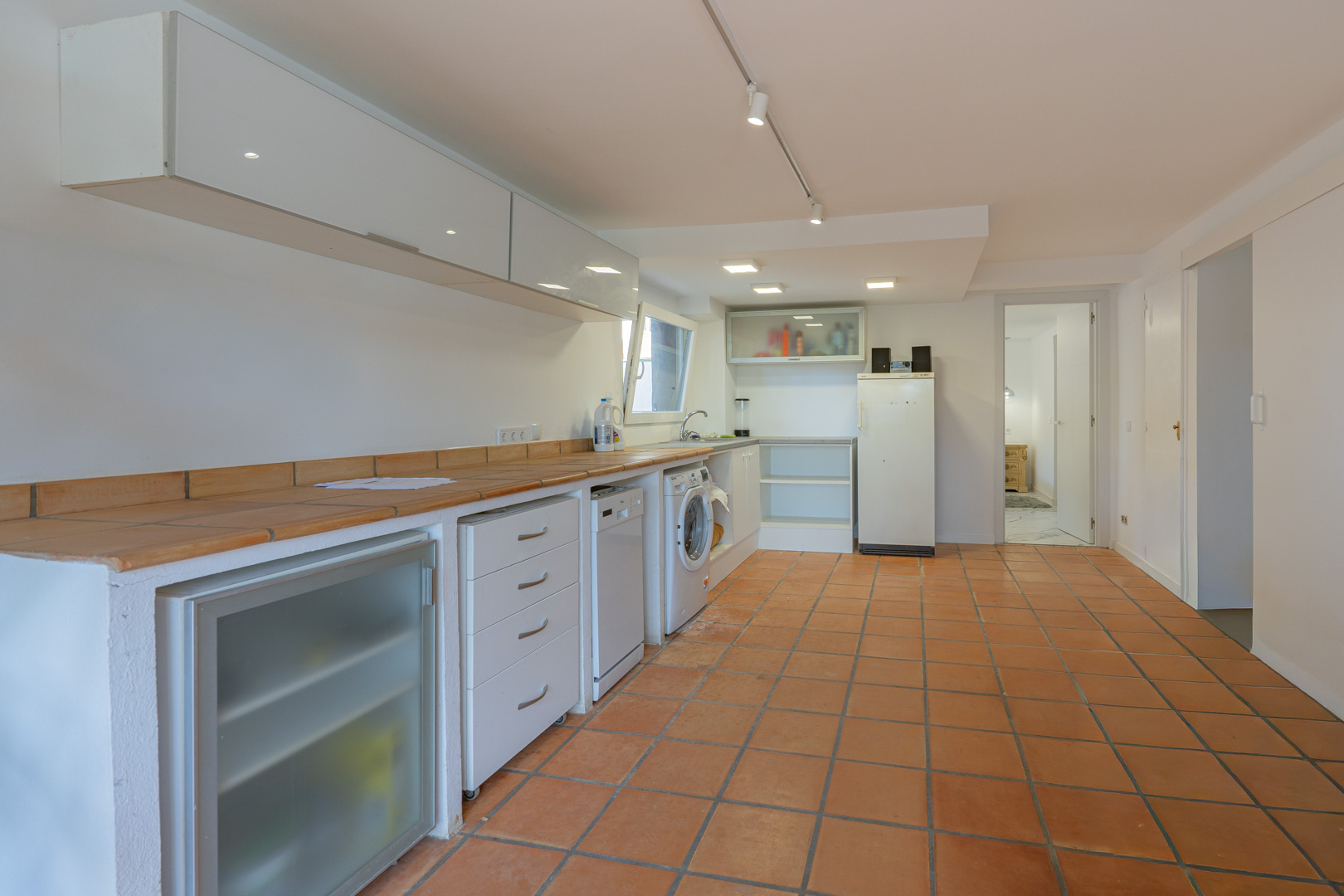 Extra keuken en wasruimte met terracotta tegelvloer, betegeld werkblad en ingebouwde apparatuur onder minimalistische witte kasten.