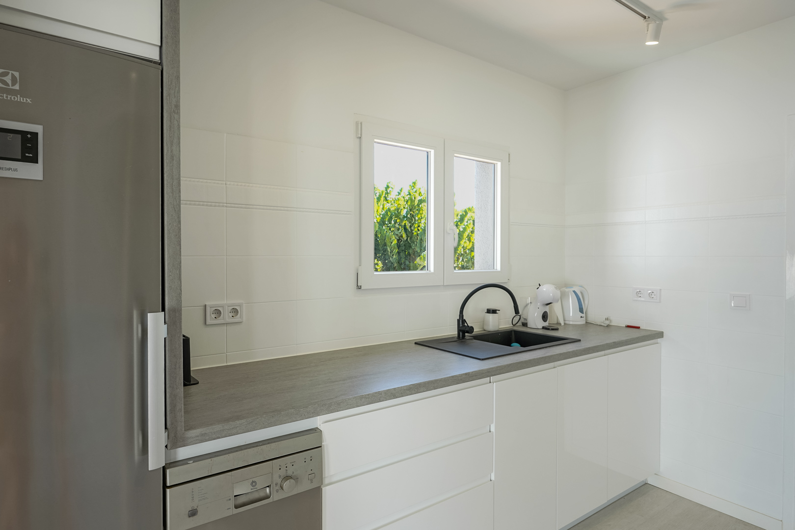 Functionele keuken met greeploze witte kasten, een grijs werkblad, zwarte spoelbak en inbouwapparatuur bij een raam met natuurlijk licht.