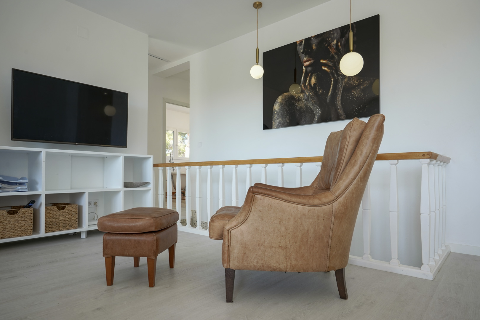 Leeshoek met een leren fauteuil en voetenbank naast de trapleuning. Voorzien van een wandtelevisie, witte open kast en moderne hanglampen.