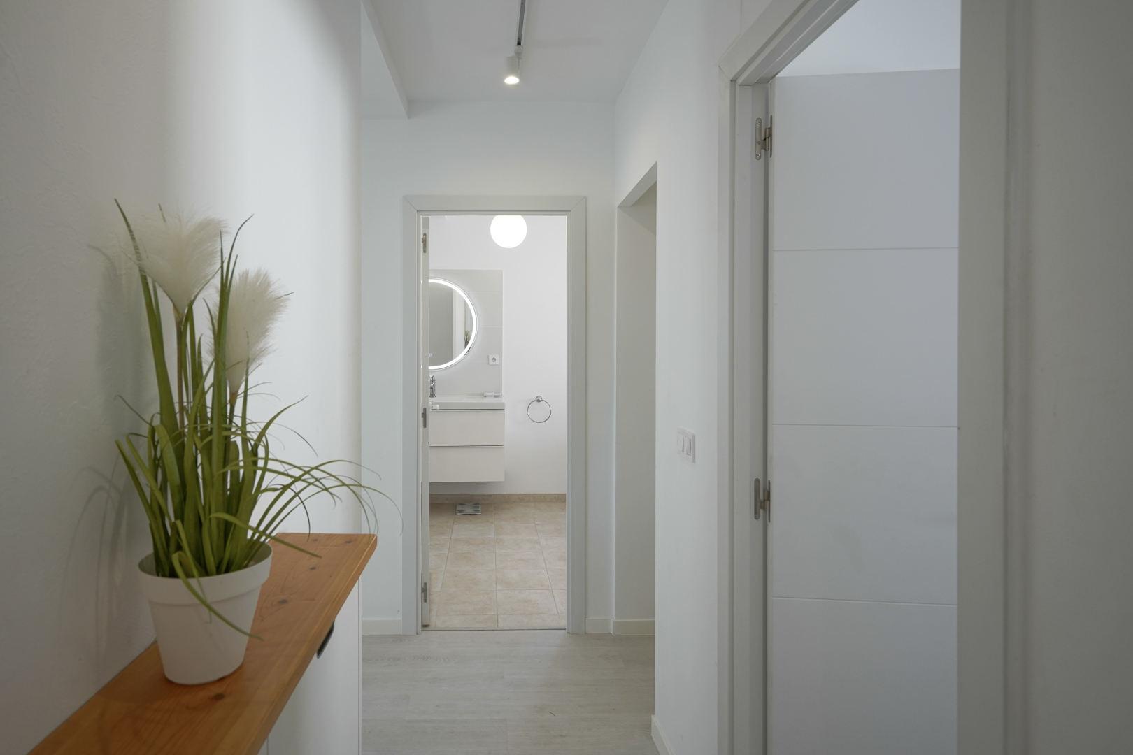 Lichte gang met witte muren en een lichte vloer die leidt naar een moderne badkamer met een verlichte ronde spiegel en zwevend wastafelmeubel.