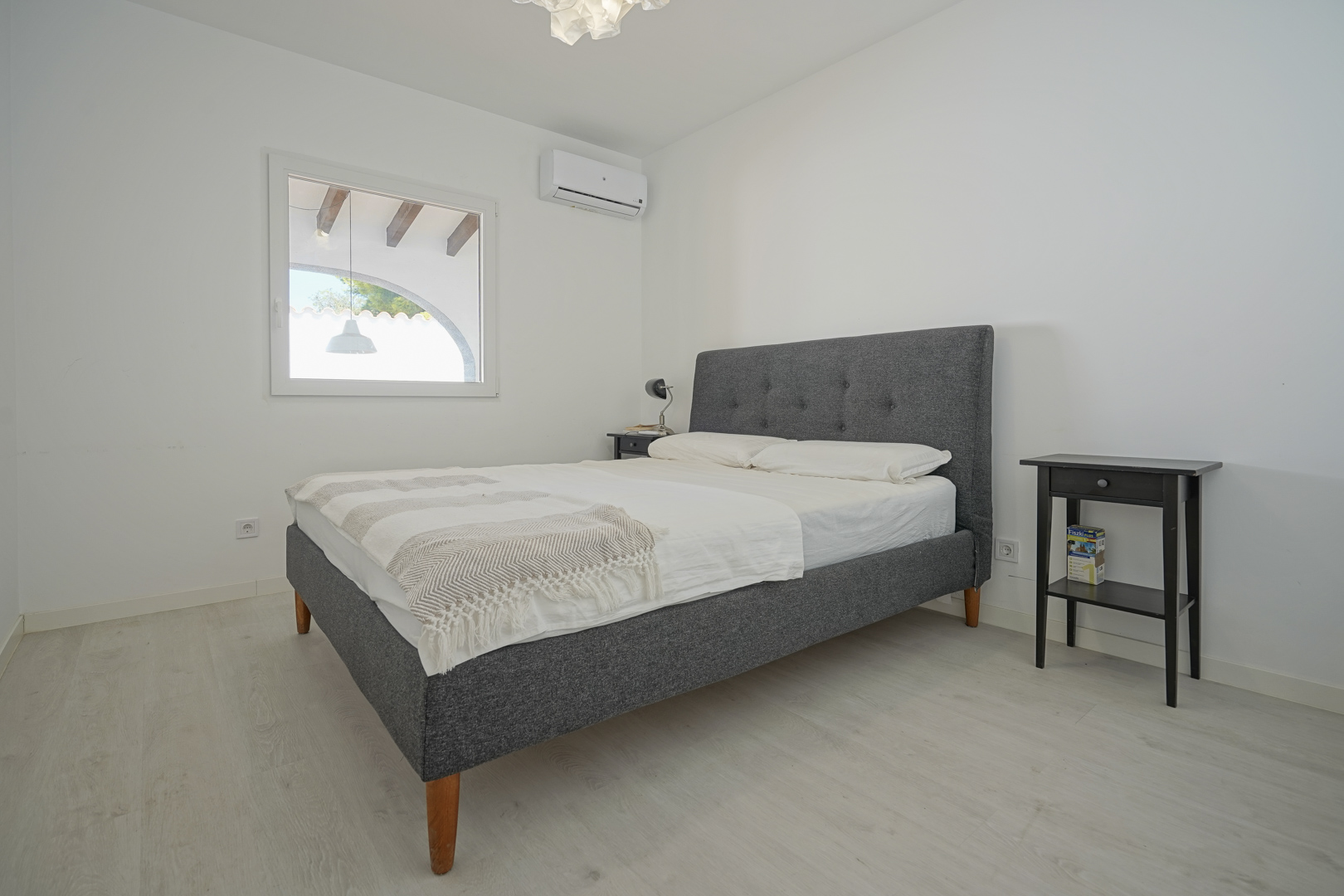 Lichte slaapkamer met grijs gestoffeerd tweepersoonsbed, airconditioning en een lichte laminaatvloer. Het raam kijkt uit op het overdekte terras met houten balken.