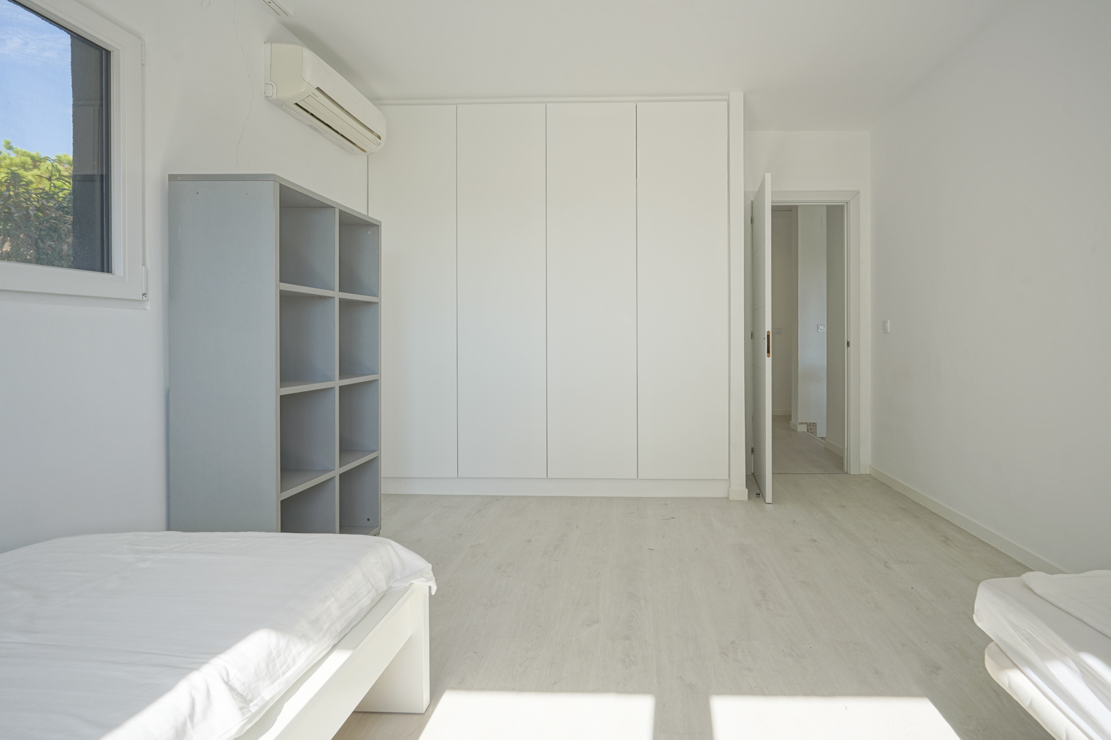 Lichte slaapkamer met kamerhoge witte inbouwkasten, lichte laminaatvloer, wandgemonteerde airconditioning en een grijze open vakkenkast.