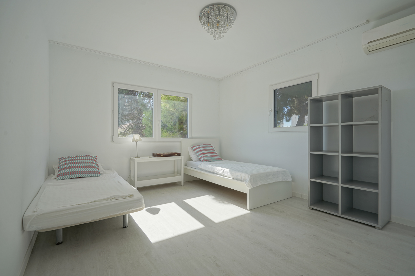 Lichte slaapkamer met twee eenpersoonsbedden, lichte houten vloer en een grijze open kast. Voorzien van airconditioning, een kristallen kroonluchter en uitzicht op bomen.