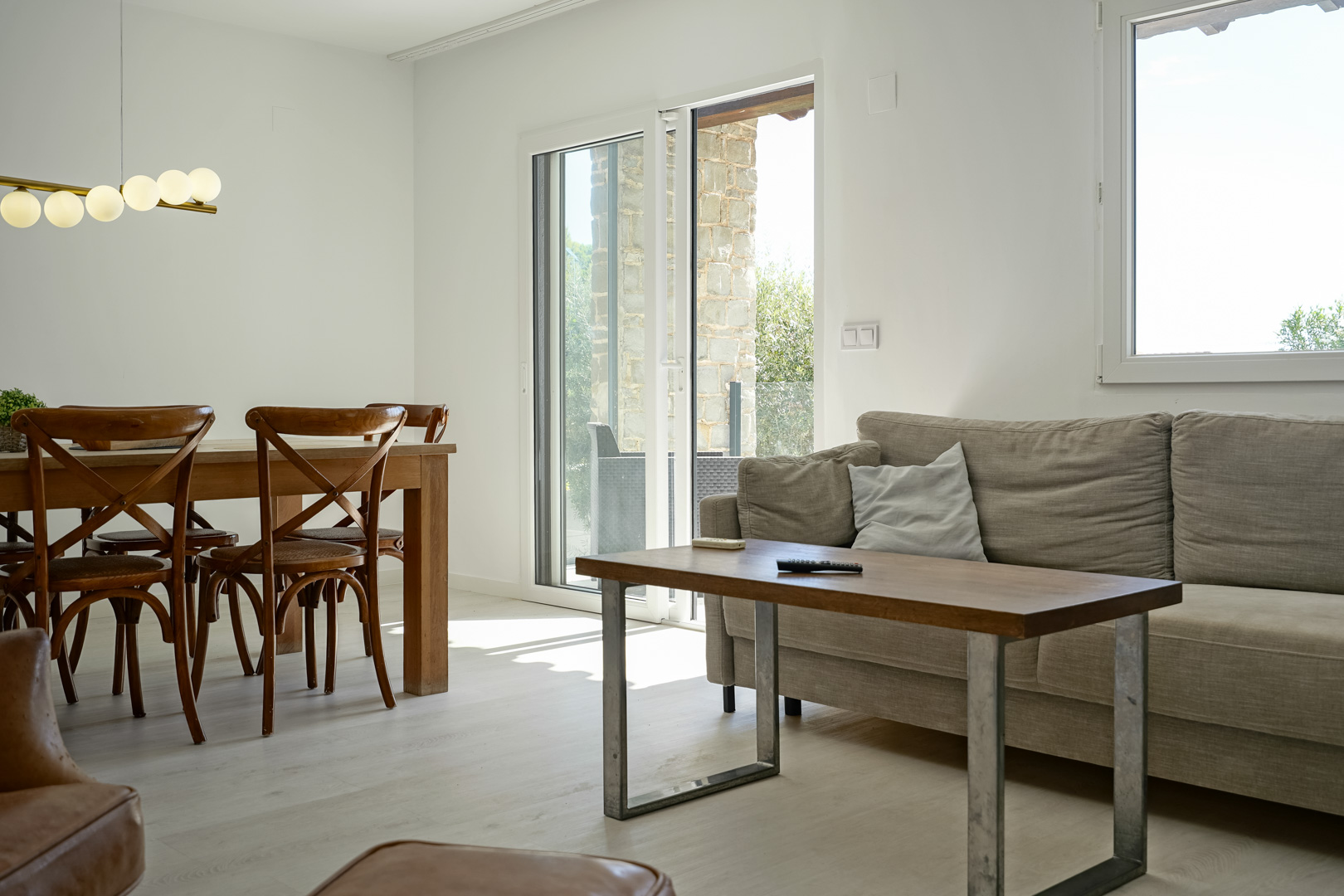 Lichte woon-eetkamer met houten meubilair en een bolvormige lamp. Deuren met dubbel glas verbinden het interieur met het natuurstenen terras.