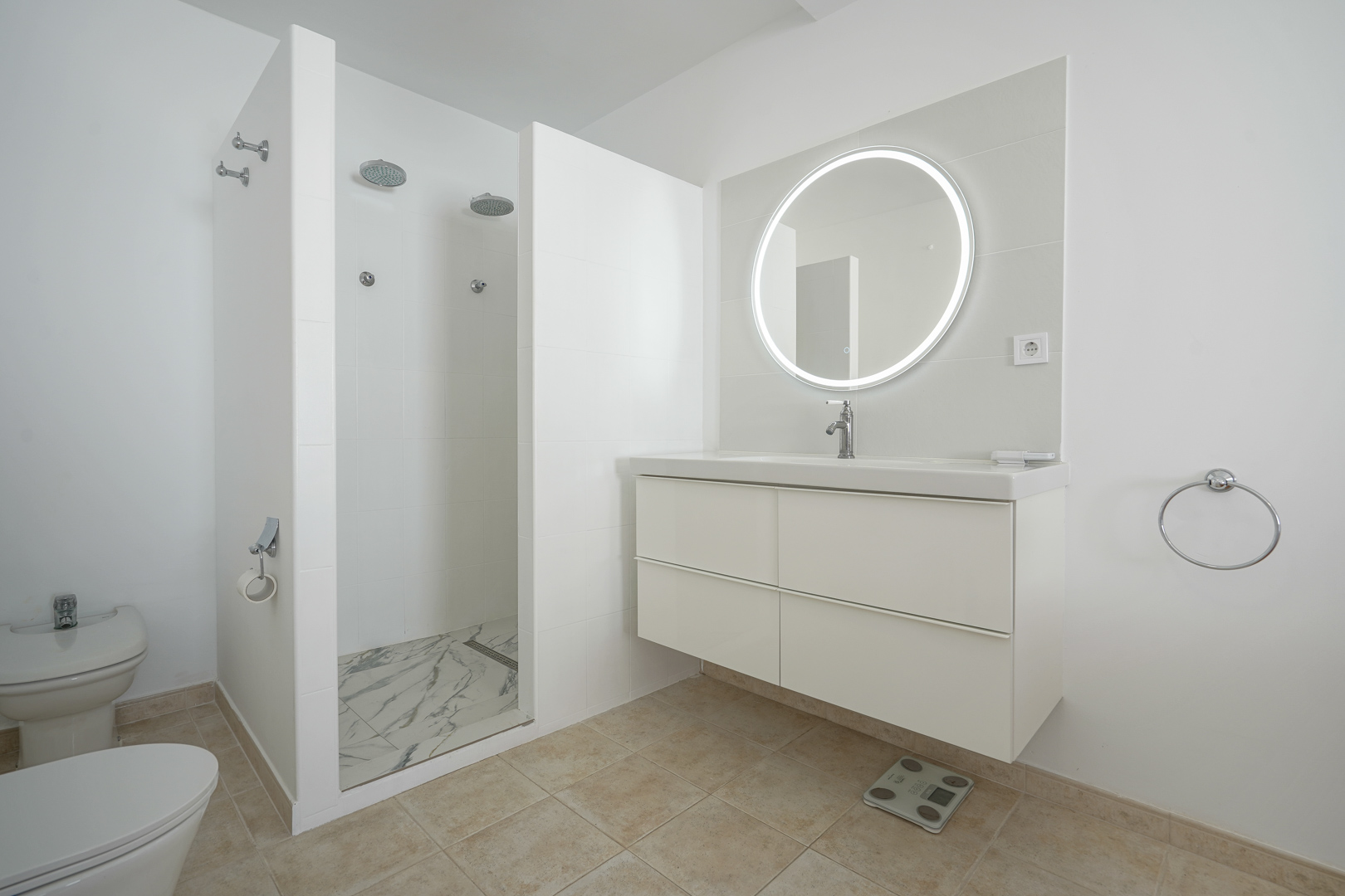 Moderne badkamer met zwevend wit wastafelmeubel, ronde verlichte spiegel en een ruime dubbele inloopdouche met vloer in marmerlook.