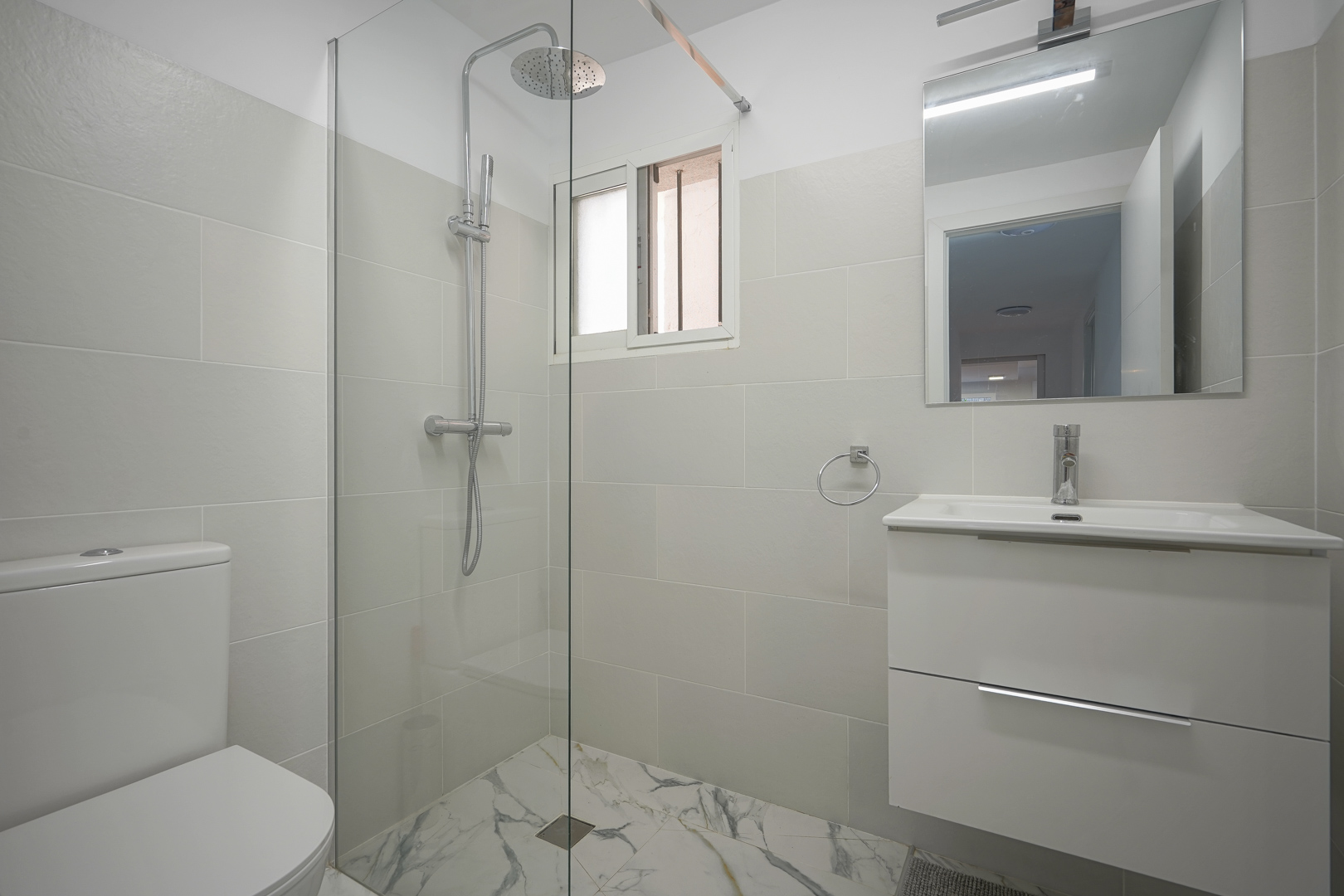 Moderne gerenoveerde badkamer met inloopdouche, glazen wand, wit wastafelmeubel en vloertegels met marmerlook.