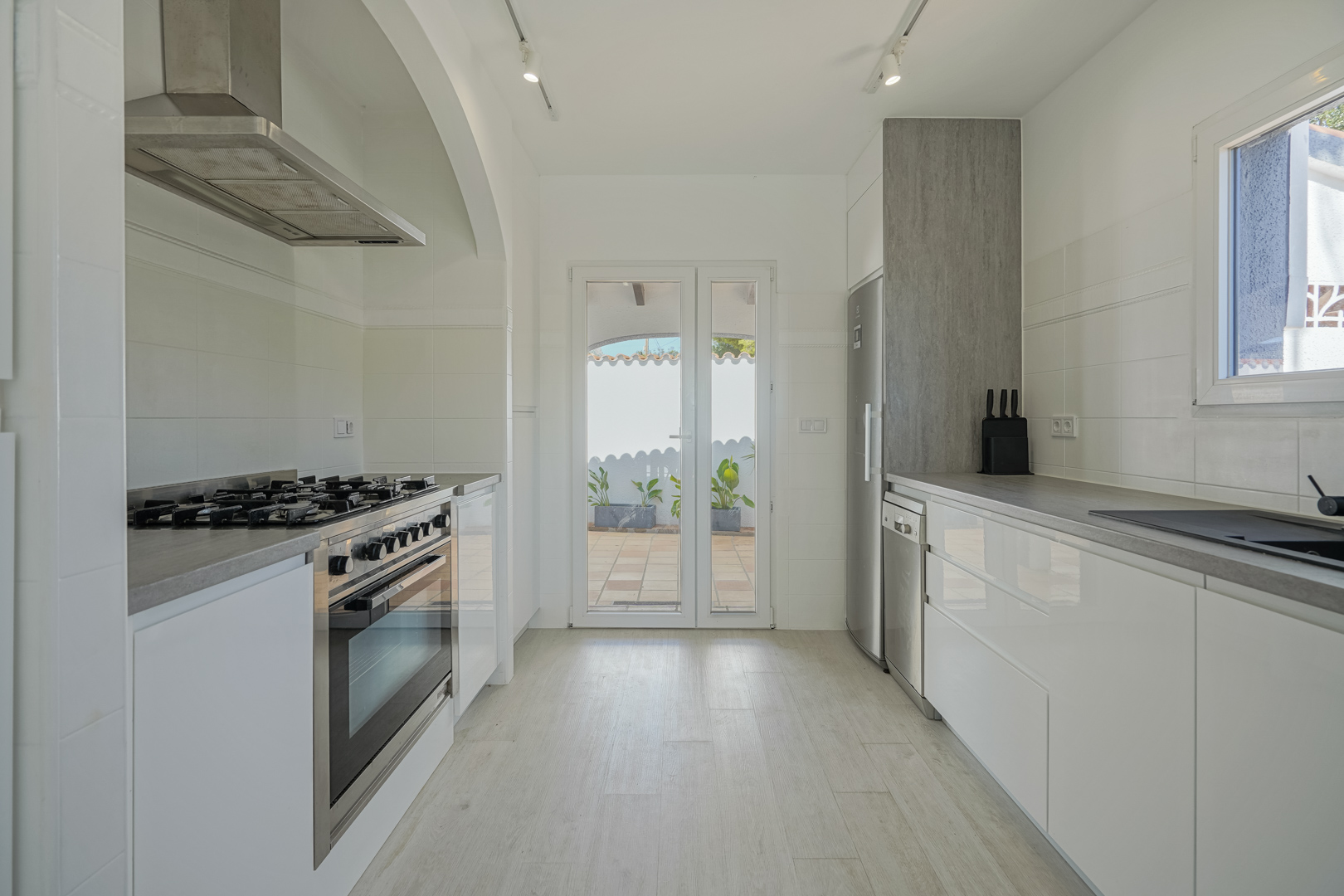 Moderne keuken met glanzend witte kasten, grijze werkbladen en rvs-apparatuur. Voorzien van een gasfornuis en directe toegang tot het buitenterras.