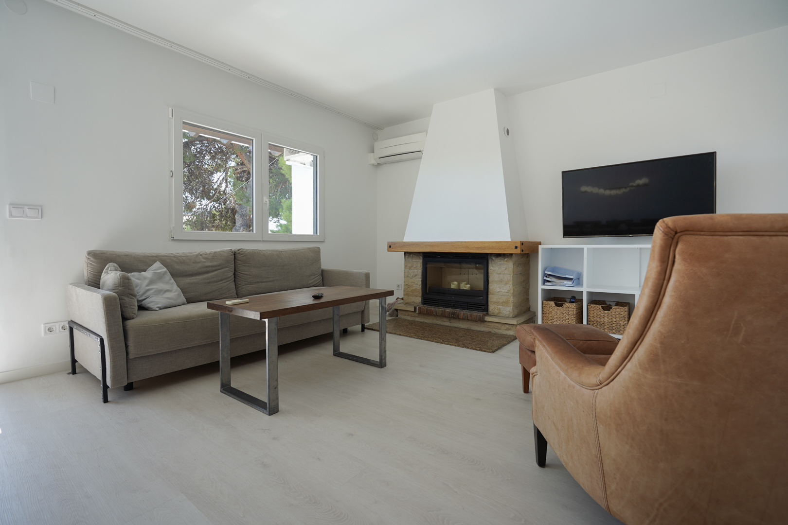 Moderne woonkamer met een stenen open haard, houten mantel, lederen fauteuil en grijze bank. Voorzien van airconditioning en dubbele beglazing voor veel lichtinval.