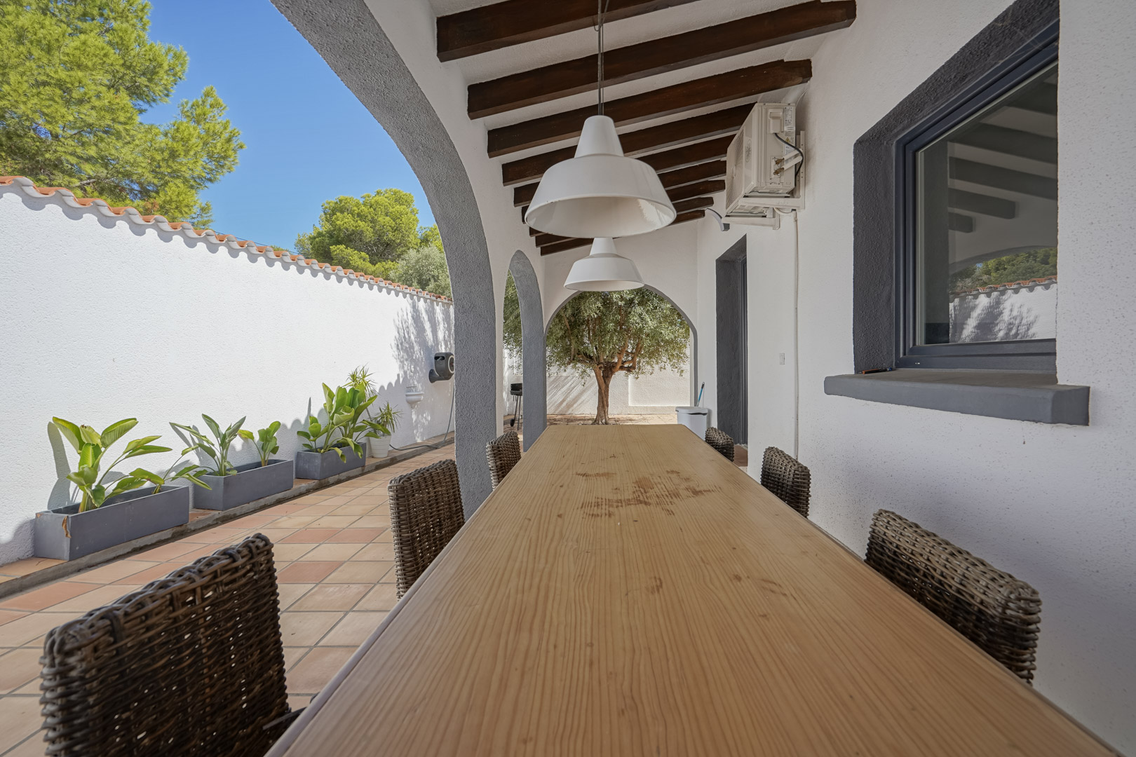 Overdekt terras met houten balkenplafond, een lange eettafel en rieten stoelen. De bogen bieden uitzicht op de mediterrane tuin met een olijfboom.