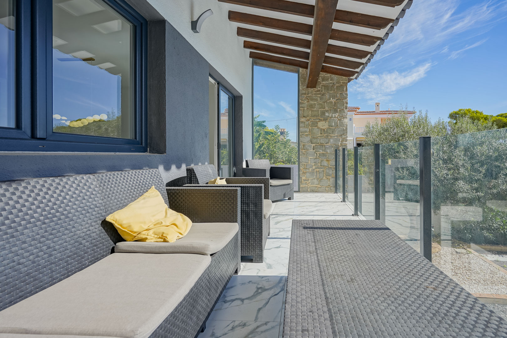 Overdekt terras met houten balkenplafond, loungeset van kunstrotan en marmeren vloer. Een glazen balustrade biedt uitzicht op de omliggende tuin.
