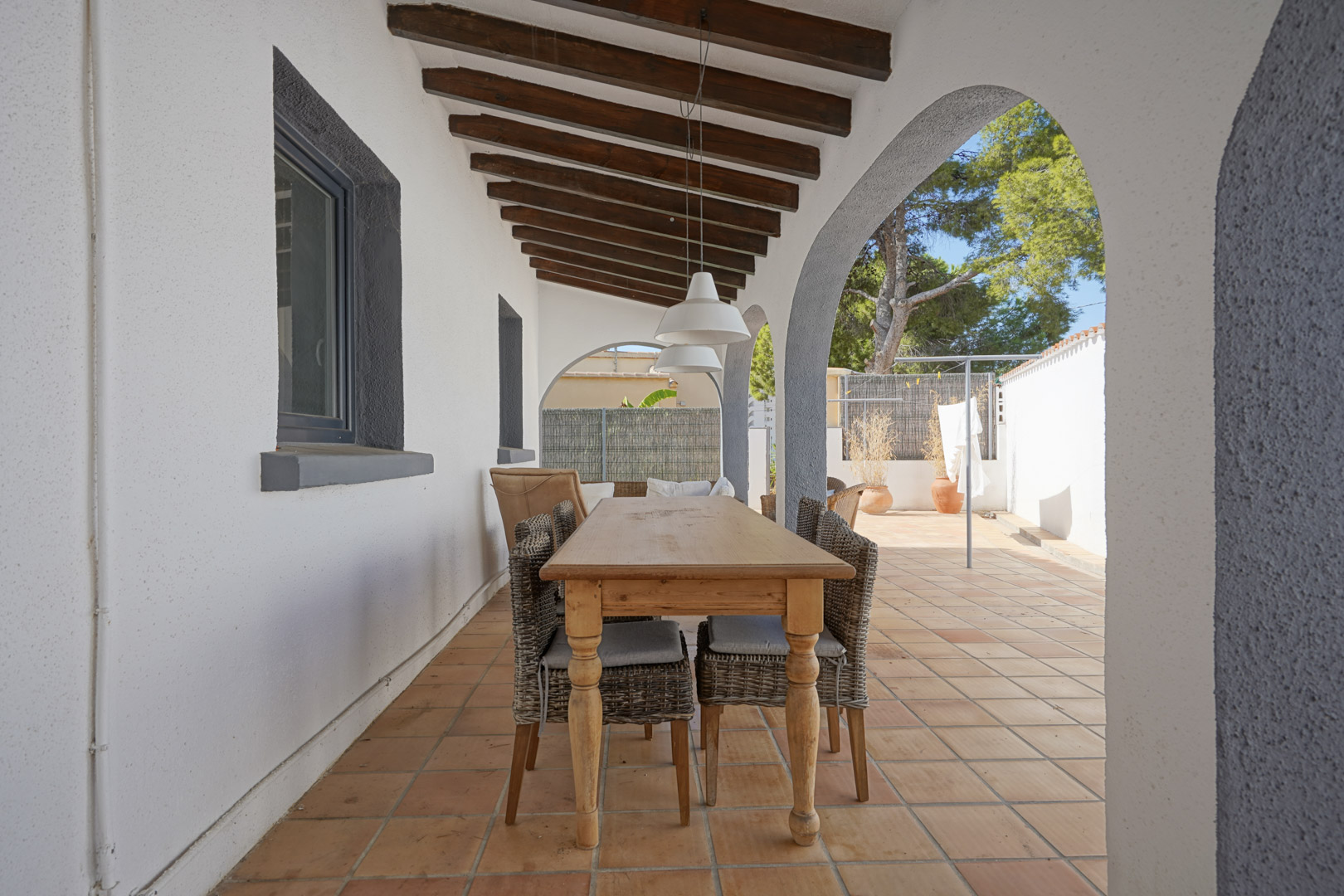 Overdekt terras met houten balkenplafond, mediterrane bogen en een eethoek buiten met houten tafel en rotan stoelen op een terracotta vloer.