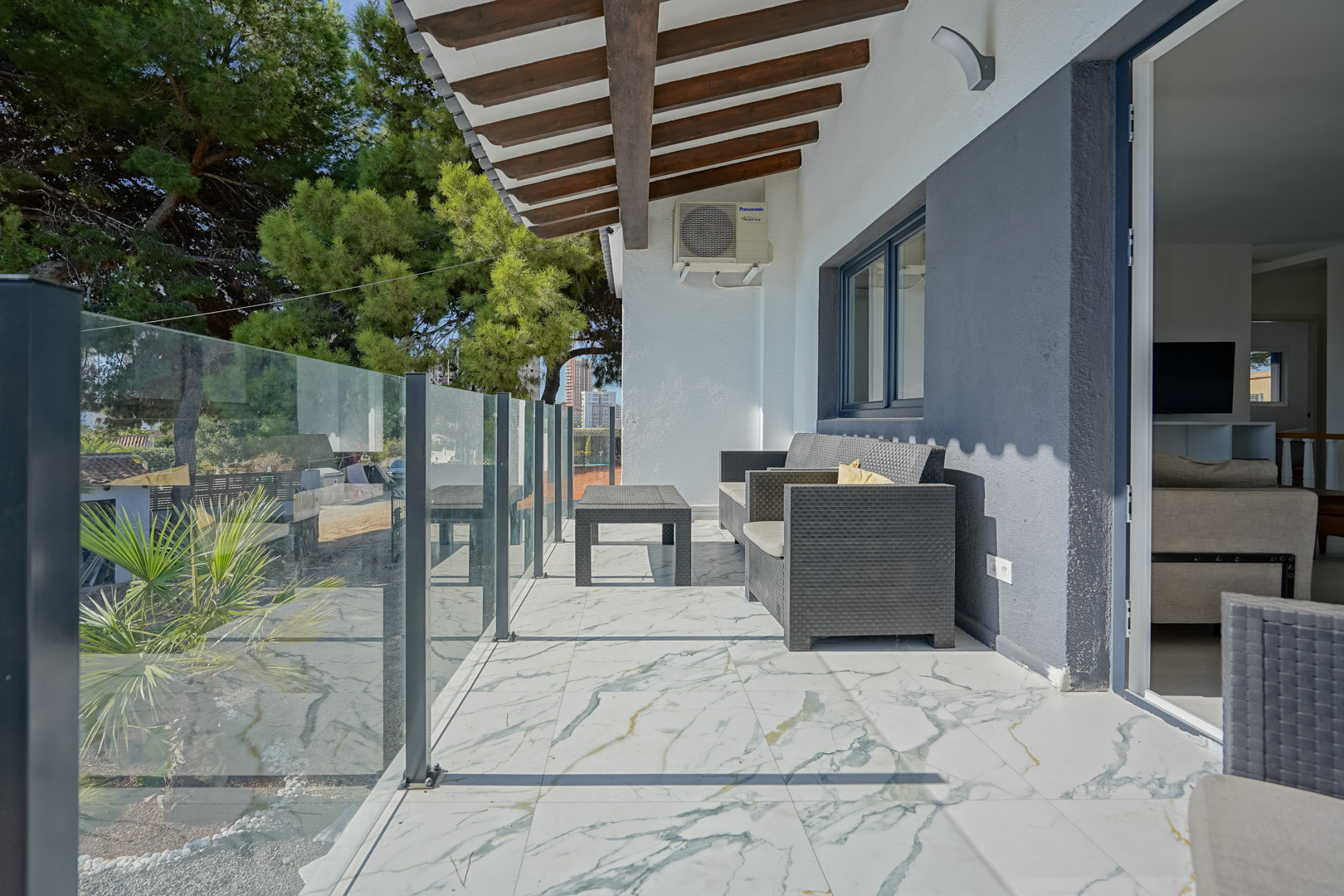 Overdekt terras met marmerlook vloertegels, een glazen balustrade en een houten balkenplafond. Voorzien van grijs rotan meubilair en uitzicht op de groene omgeving.