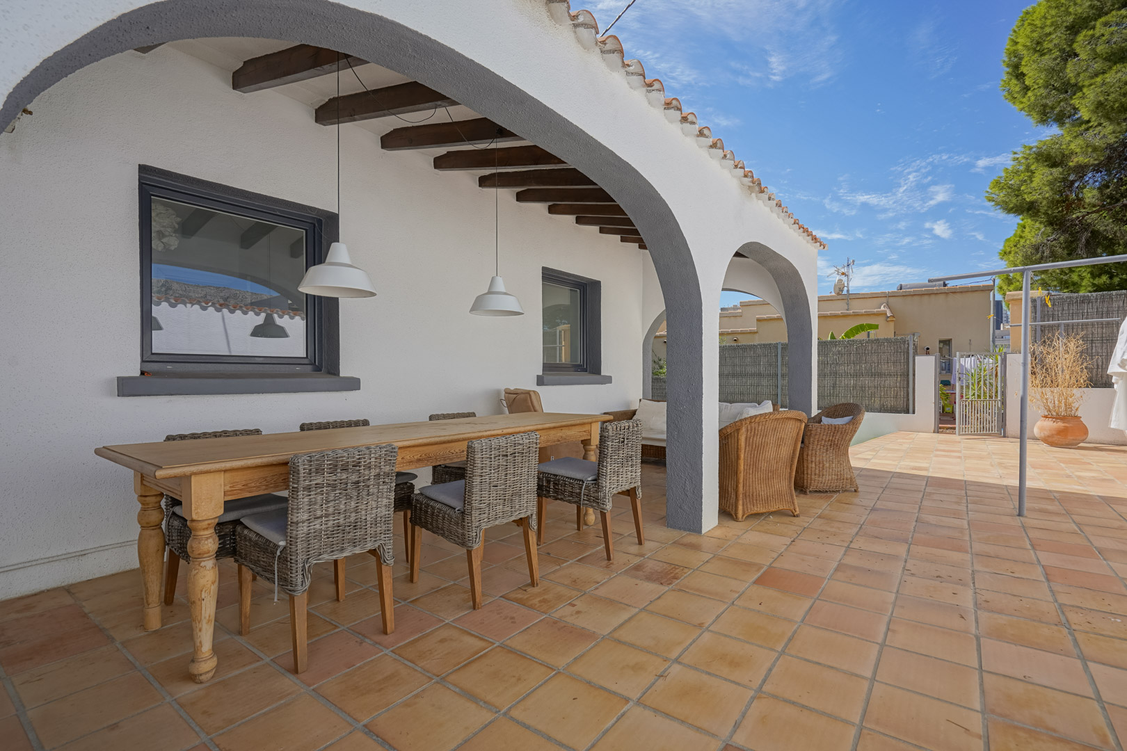 Overdekt terras met witte bogen, houten balken en terracotta vloer. Ingericht met een houten eettafel en rotan stoelen onder moderne hanglampen.