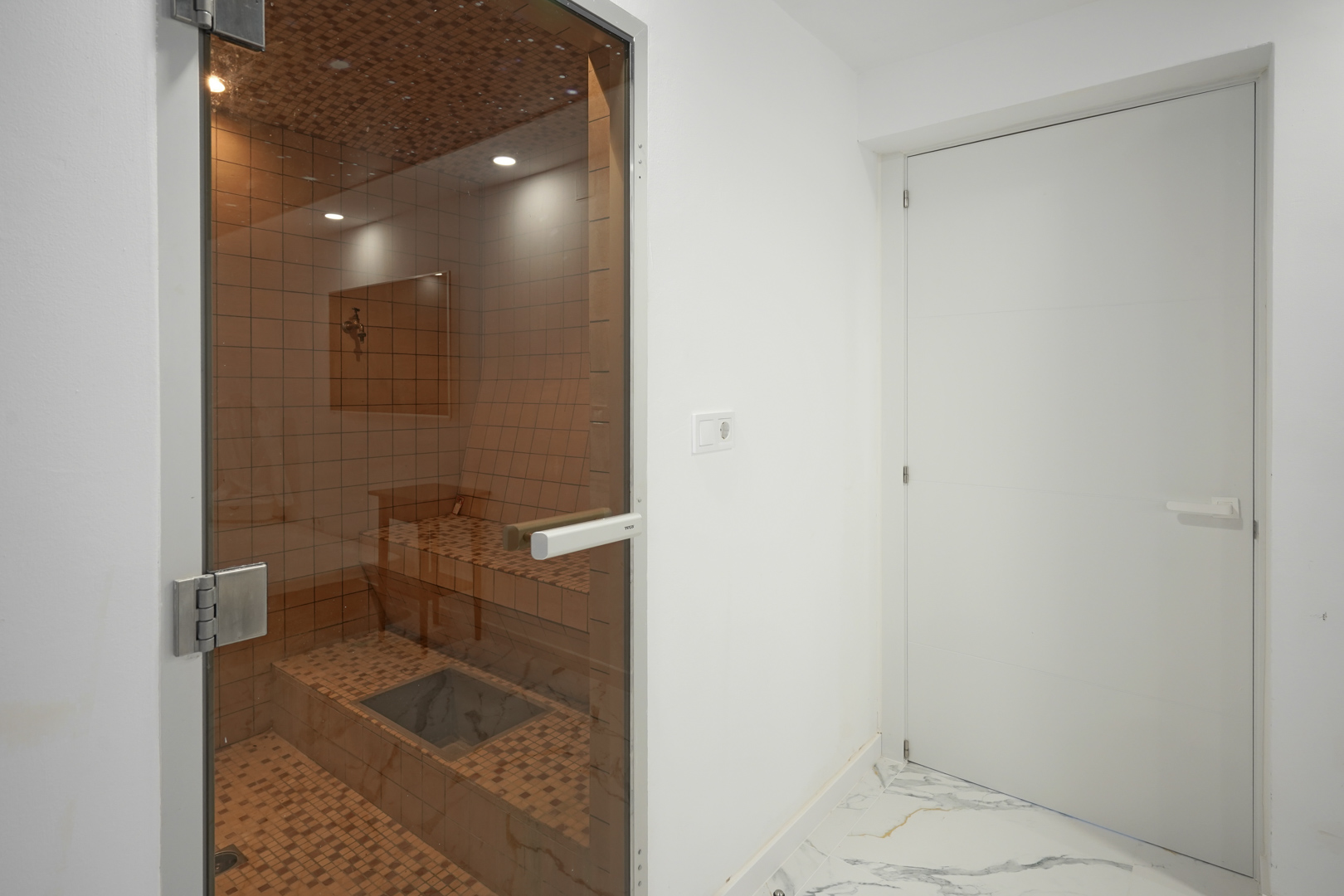 Privésauna met keramische mozaïektegels, inbouwspots en een glazen deur, gelegen naast een ruimte met een wit marmeren vloer.