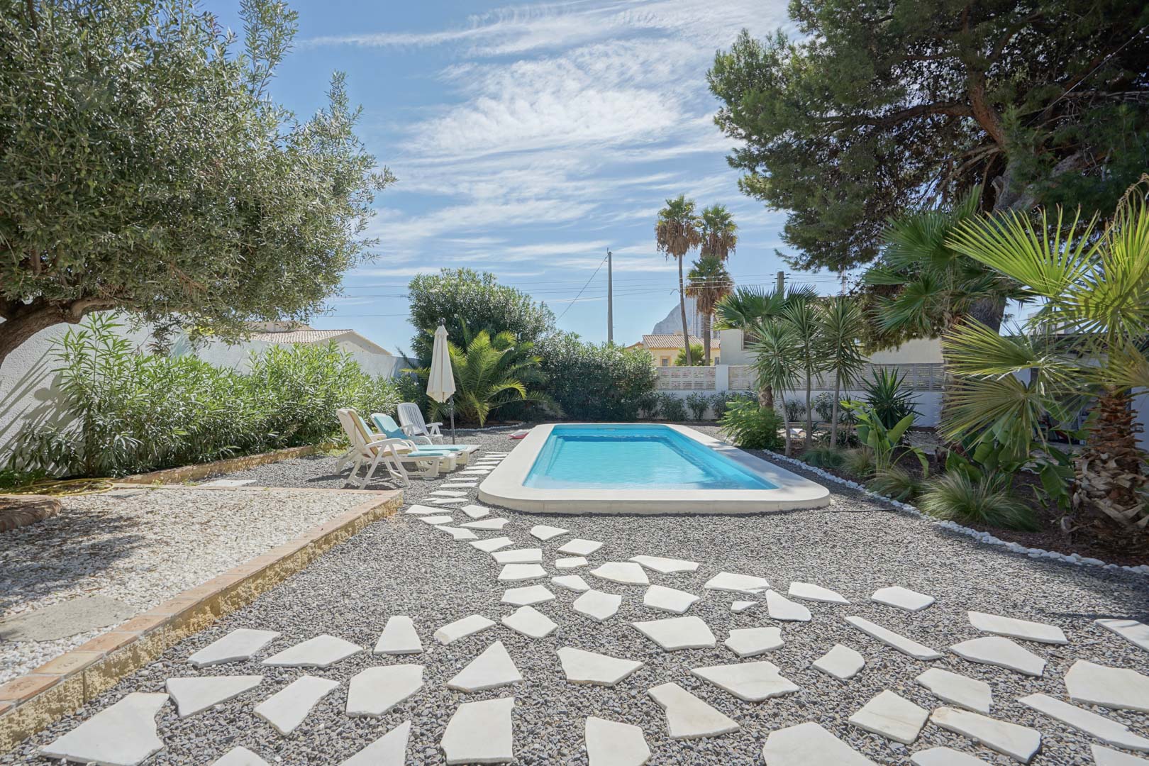 Rechthoekig zwembad in een mediterrane tuin met olijfbomen en palmen. Voorzien van een pad van onregelmatige stenen op grind en een zonneterras.