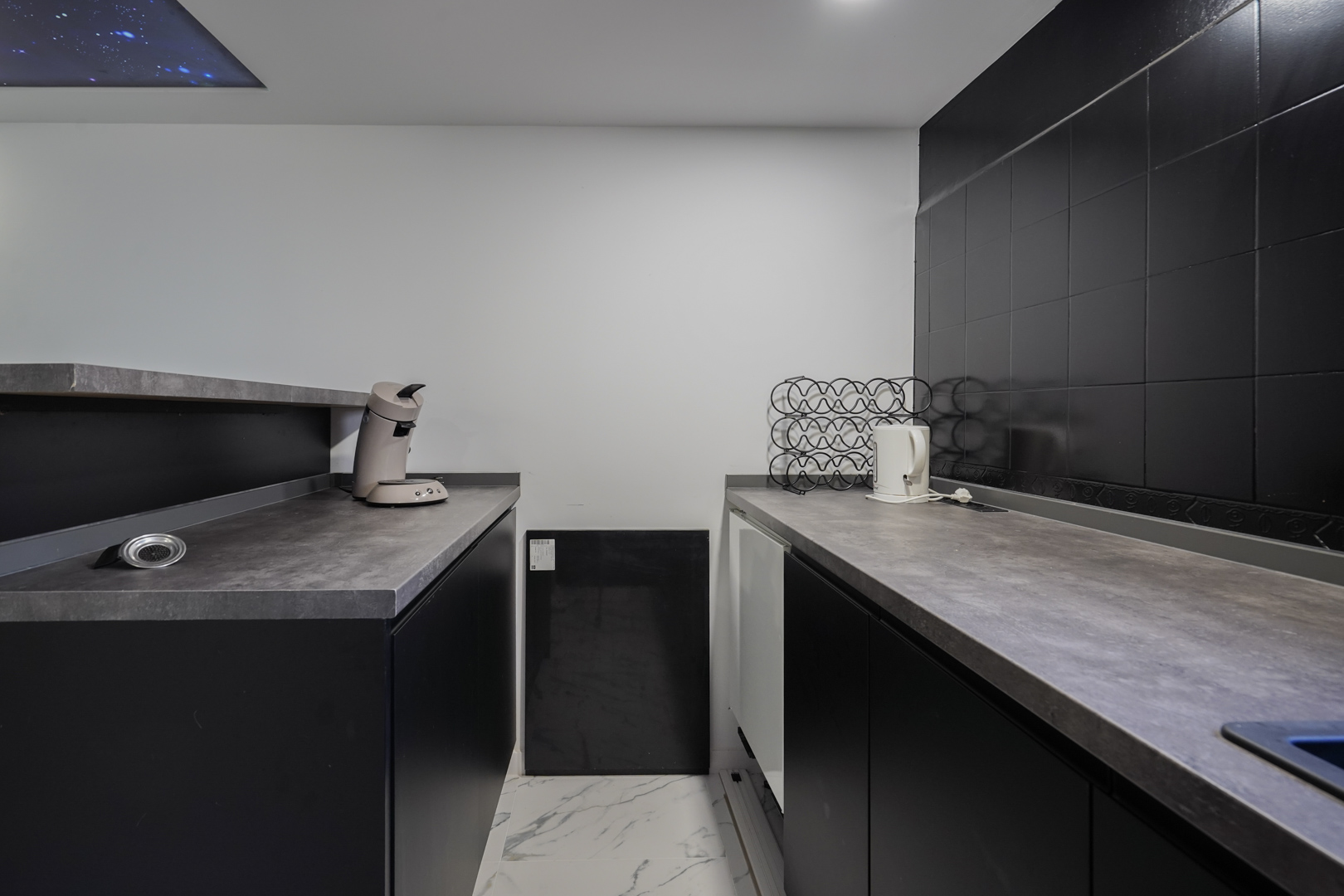 Secundaire keuken met zwarte kasten, grijze werkbladen en een marmerlook vloer. Voorzien van een decoratief plafondpaneel met sterrenhemel-effect.