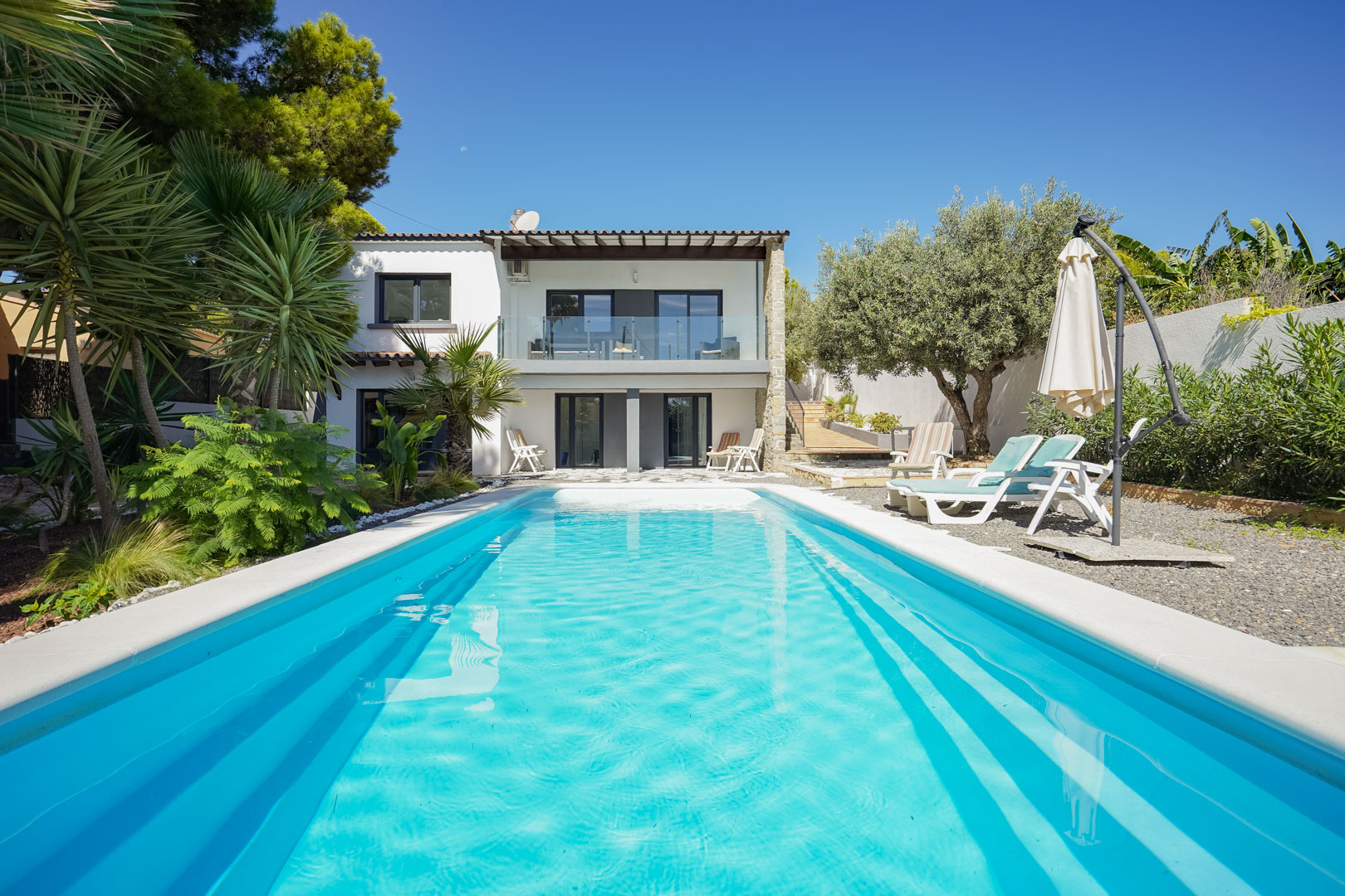 Uitzicht op het rechthoekige zwembad en de mediterrane tuin met palmbomen, met de witte gevel van de villa en het boventerras met glazen balustrade.