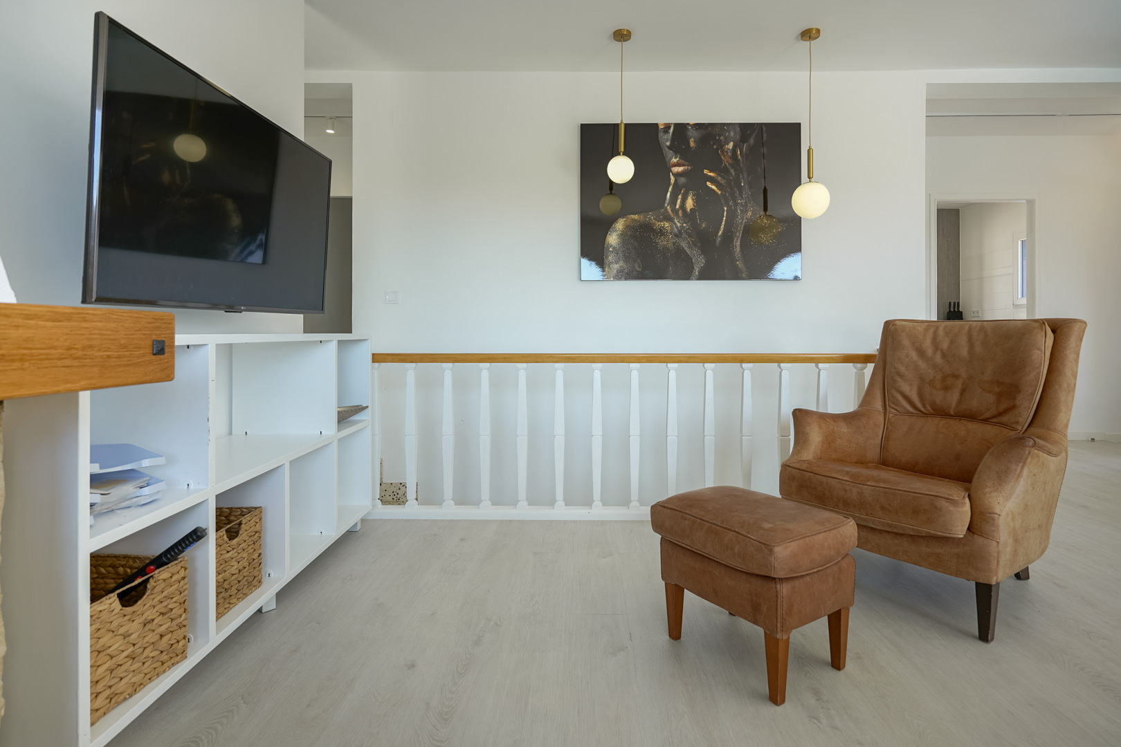 Zithoek met lederen fauteuil en voetenbank, witte open kast en houten balustrade. Voorzien van moderne hanglampen en een lichte laminaatvloer.