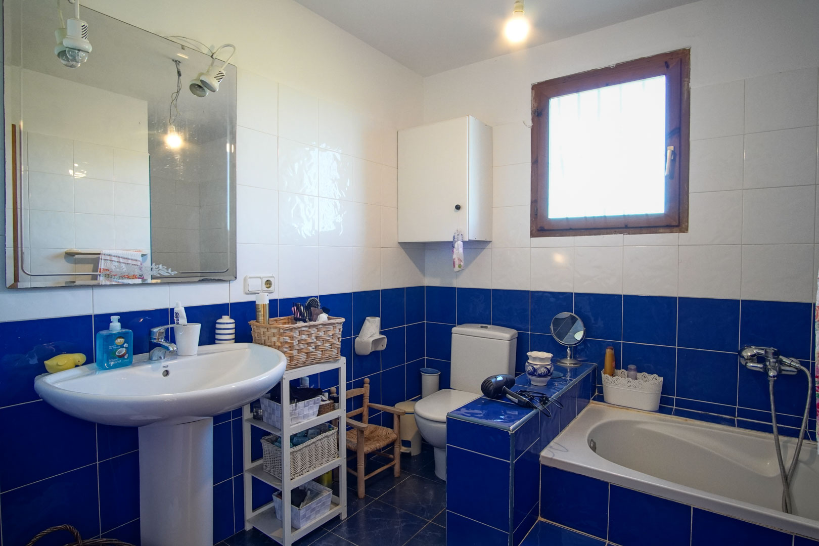 Badkamer met blauw-witte betegeling, voorzien van een kolomwastafel, inbouwbad en een houten raamkozijn voor natuurlijk lichtinval.