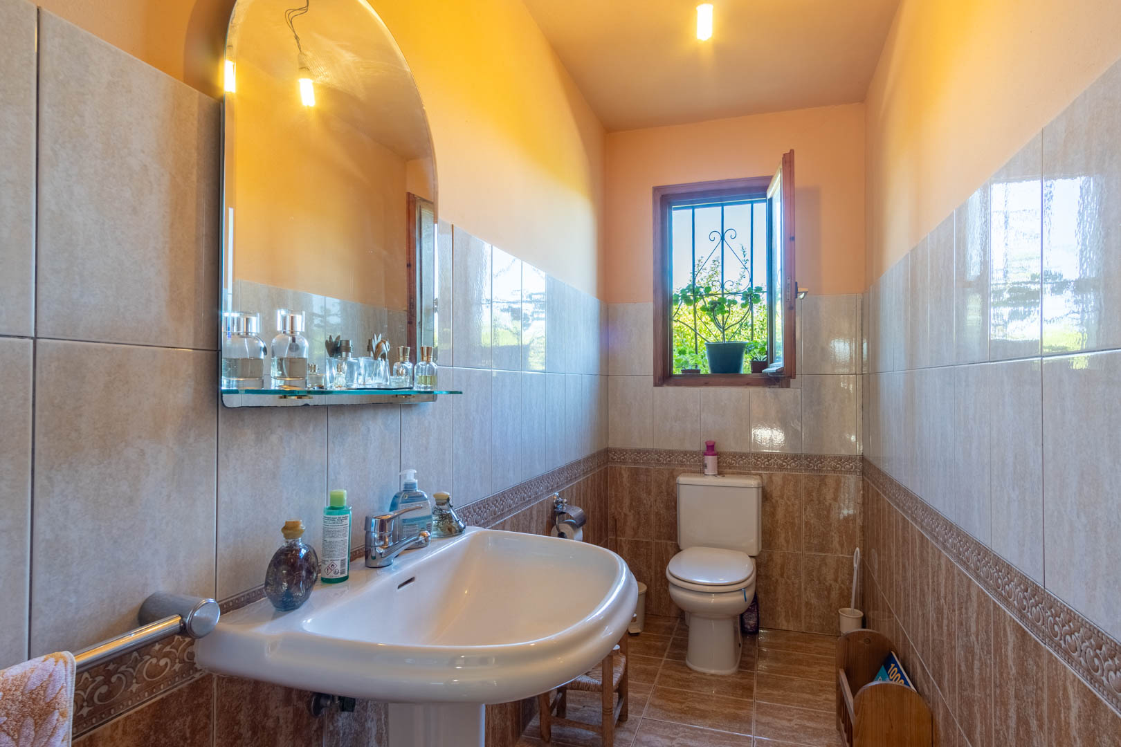 Badkamer met wandtegels in aardetinten, een kolomwastafel, een boogspiegel en een houten raam met natuurlijk licht.