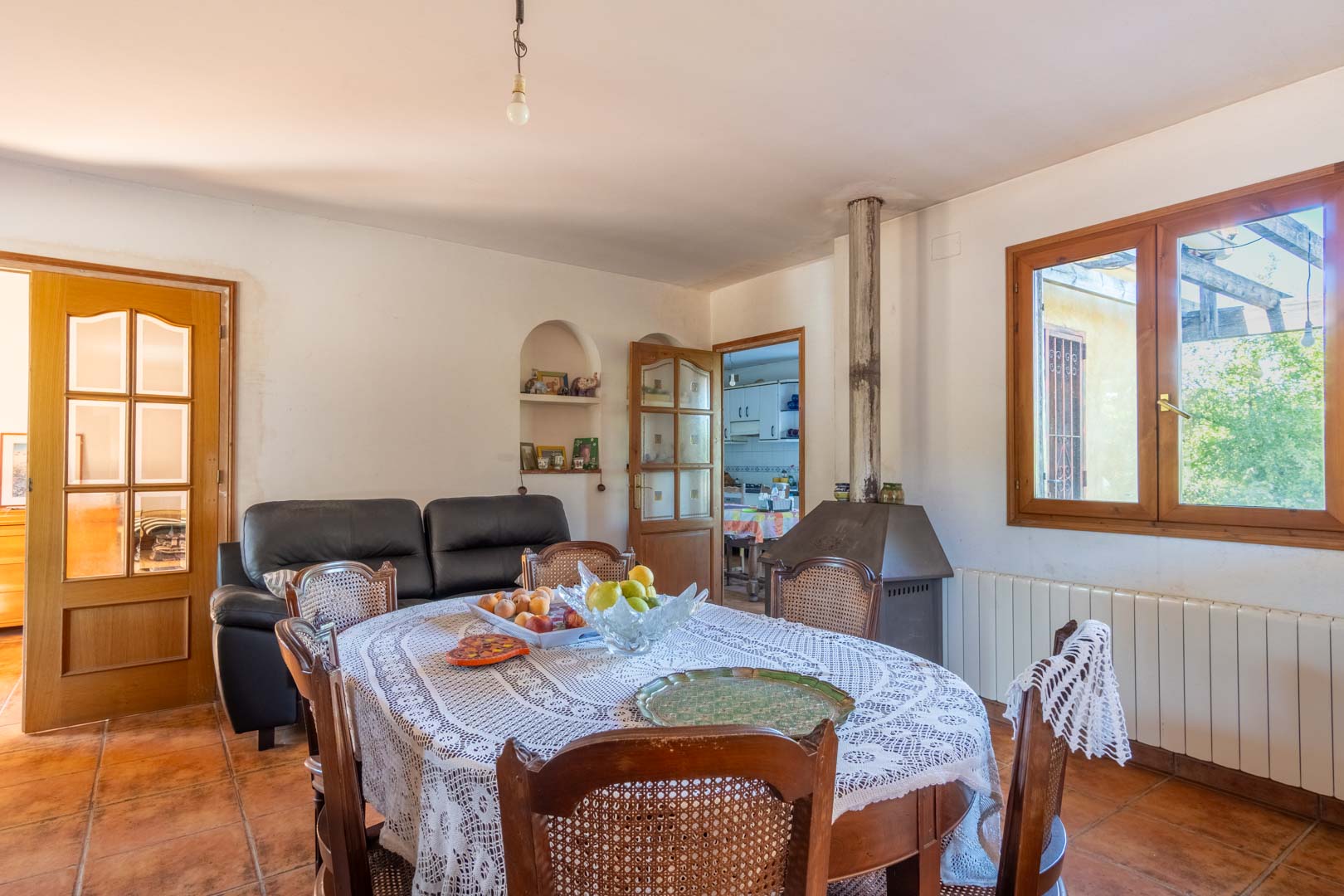 Eetkamer met terracotta vloertegels, een houtkachel en klassiek houten meubilair. De ruimte geniet van natuurlijk licht via het houten kozijn en biedt toegang tot de keuken.