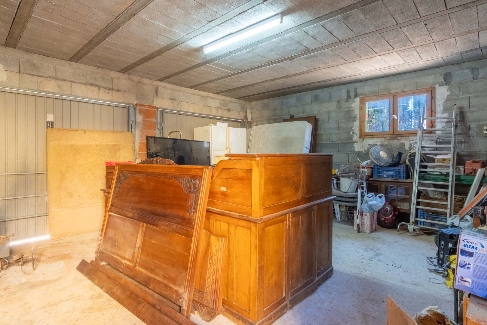 Ruime opslagruimte of garage met wanden van betonblokken, balkenplafond, TL-verlichting en een houten raam voor natuurlijk lichtinval.