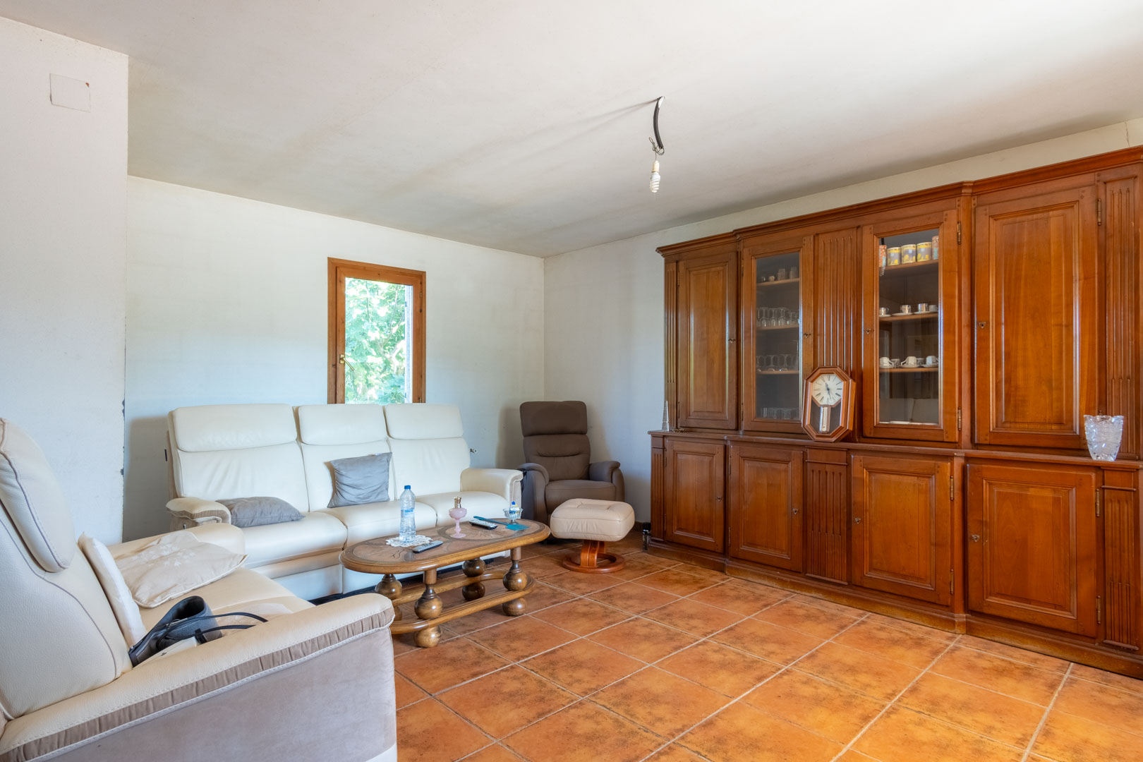 Woonkamer met terracotta tegelvloer, witte lederen banken en een grote massief houten wandkast voorzien van glazen vitrines.