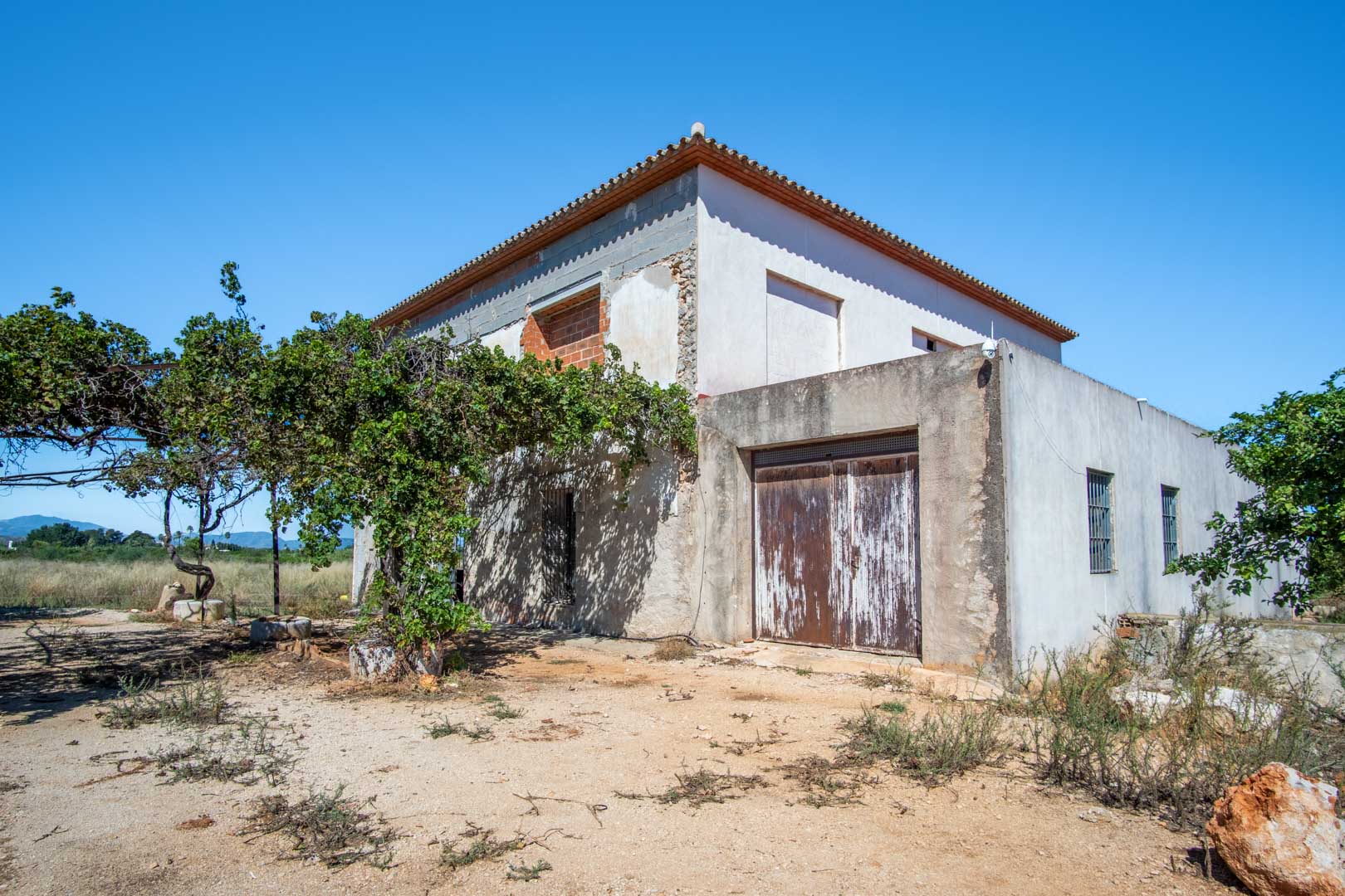 Buitenaanzicht van het landhuis in renovatie, met een solide basisstructuur, vlak terrein en vrij uitzicht op de nabijgelegen bergen.