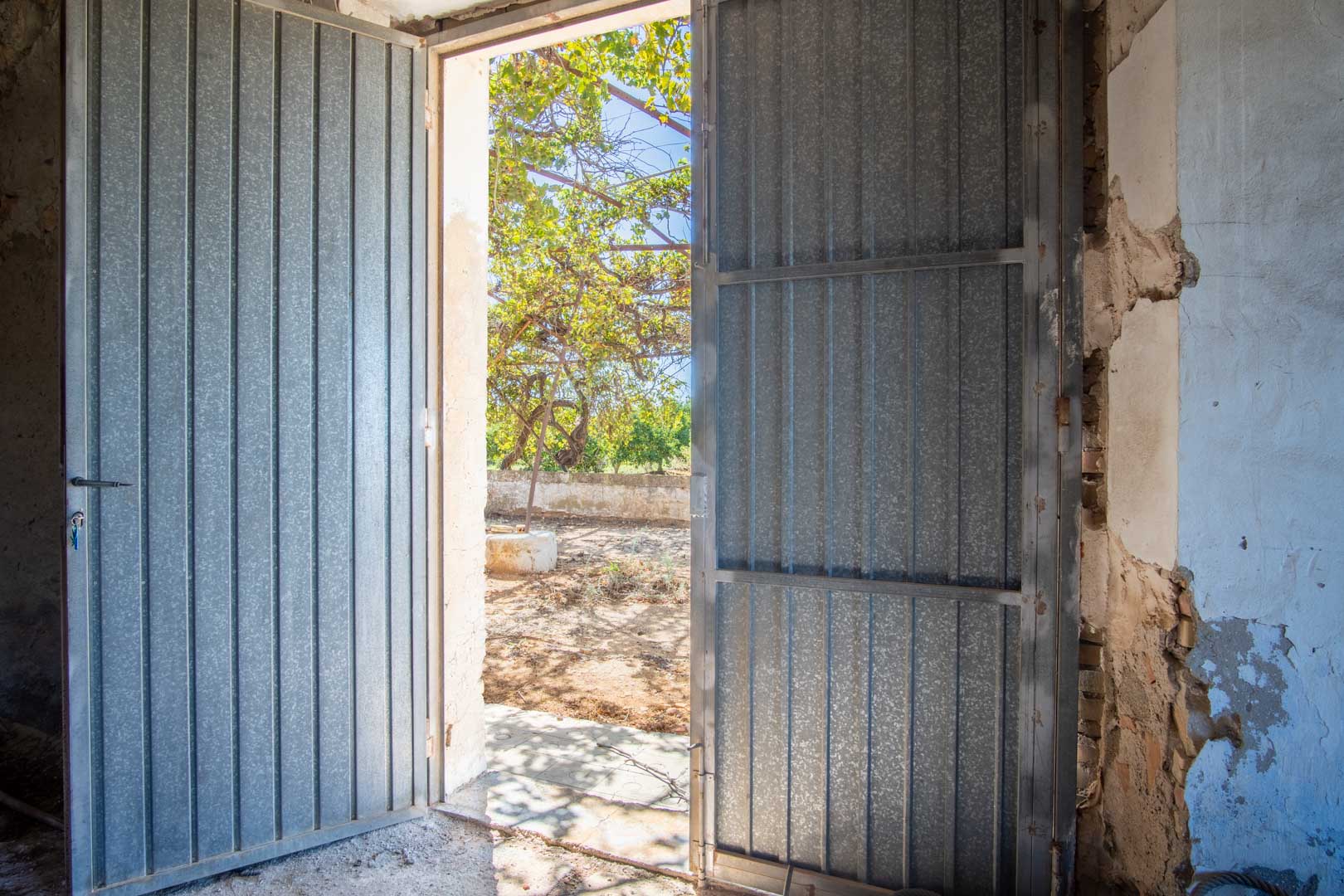 Hoofdtoegang via grote metalen deuren die het open interieur verbinden met het buitenperceel en de mediterrane vegetatie.