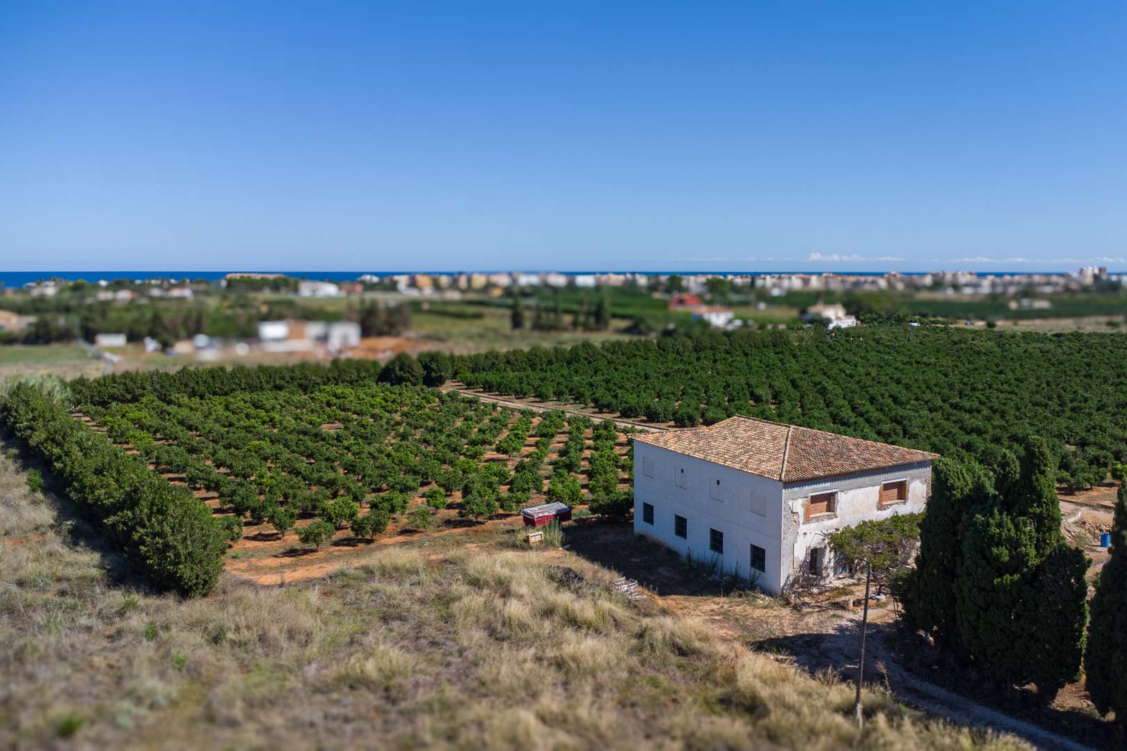 Luchtfoto van het landhuis omgeven door boomgaarden, met een witte gevel, pannendak en panoramisch uitzicht op de Middellandse Zee aan de horizon.