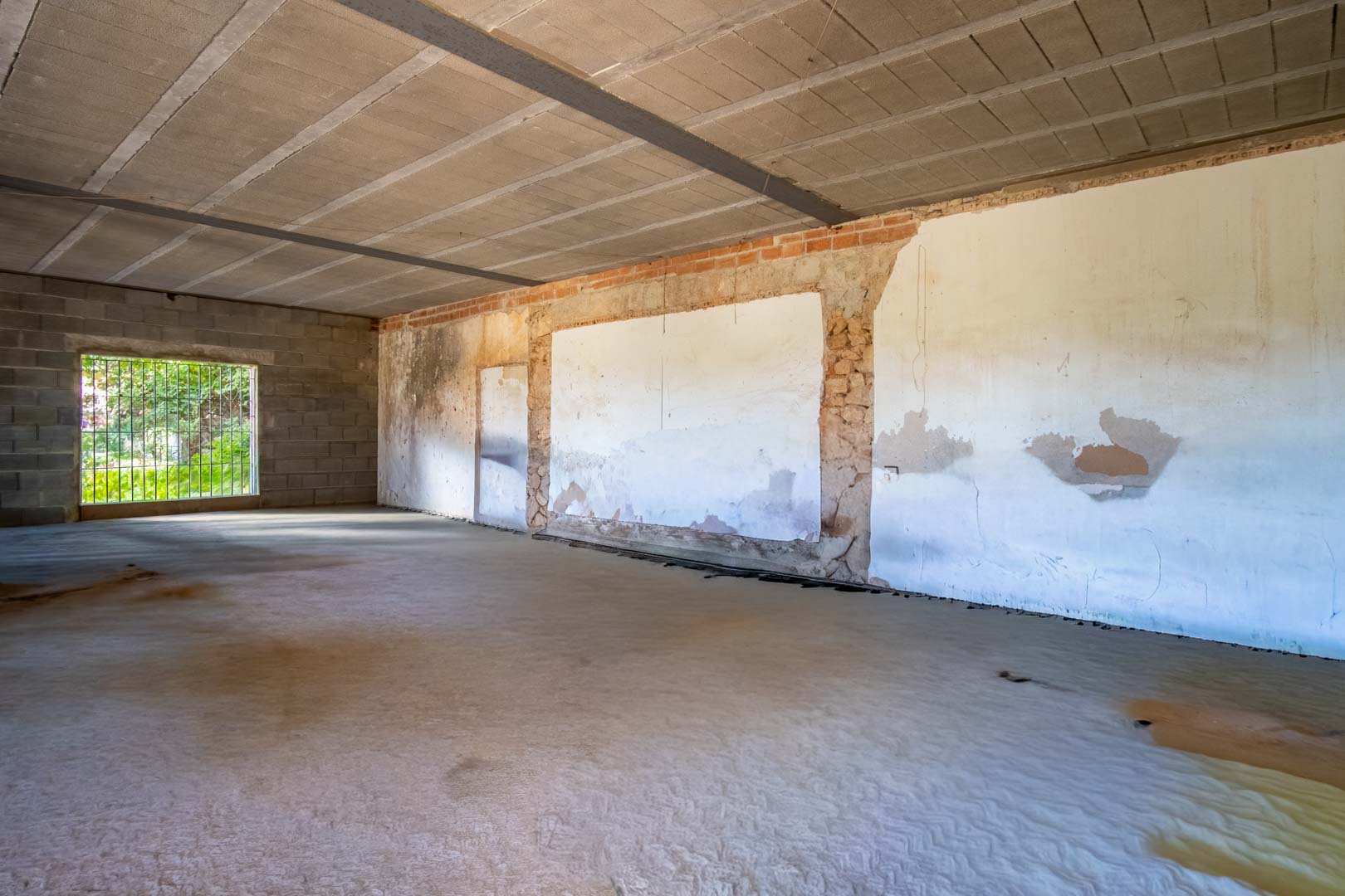 Open binnenruimte in renovatiefase met betonnen vloer, zichtbare plafondbalken en muren van een combinatie van natuursteen en baksteen.