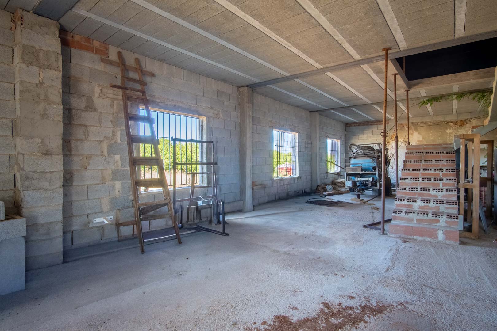 Ruime, open binnenruimte in renovatie met muren van betonblokken, een balkenplafond en ramen met tralies die zorgen voor natuurlijk licht en uitzicht.