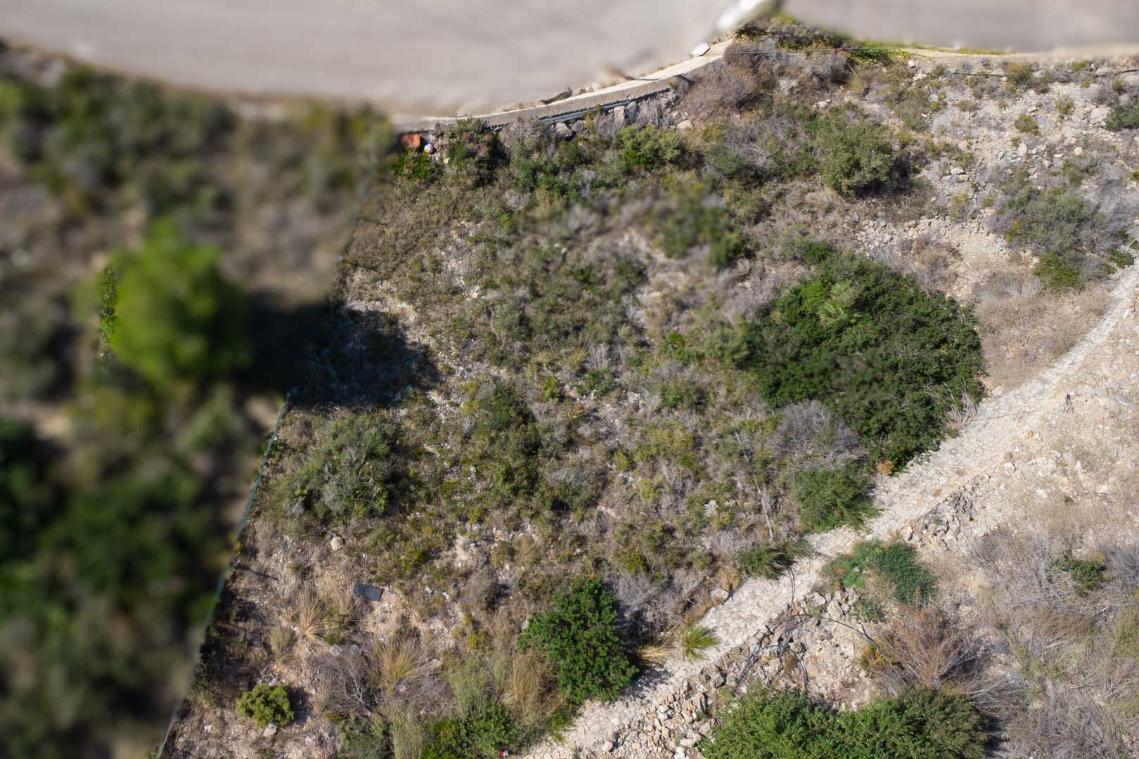 Luchtfoto van het hellende bouwperceel met inheemse mediterrane begroeiing, rotsachtige bodem en een stenen begrenzingpad.