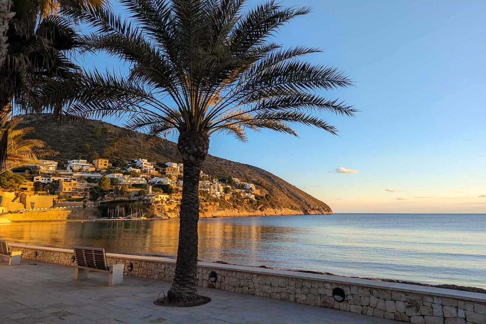 Met steen geplaveide promenade in El Portet met palmbomen en bankjes, uitkijkend over de rustige baai en de villa's op de heuvel tijdens het gouden uur.