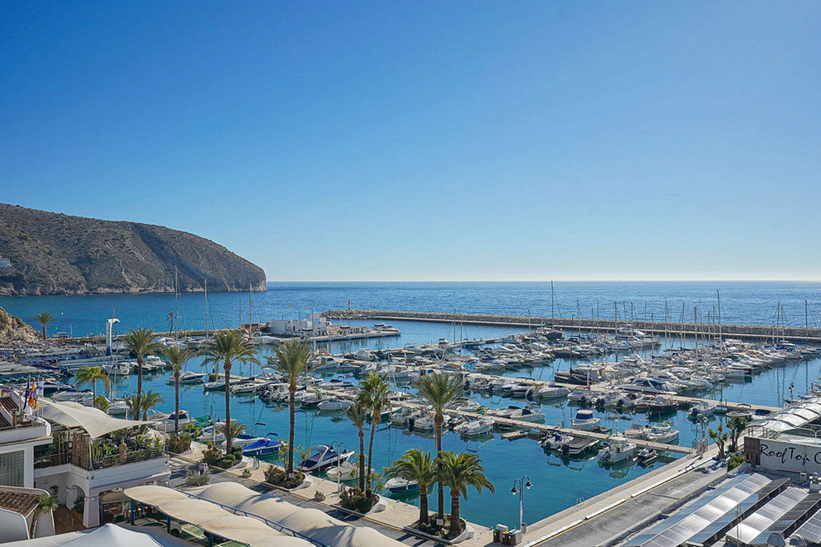 Panoramisch uitzicht op de jachthaven van Moraira met aangemeerde boten, palmbomen en de kustlijn tegen een helderblauwe lucht.