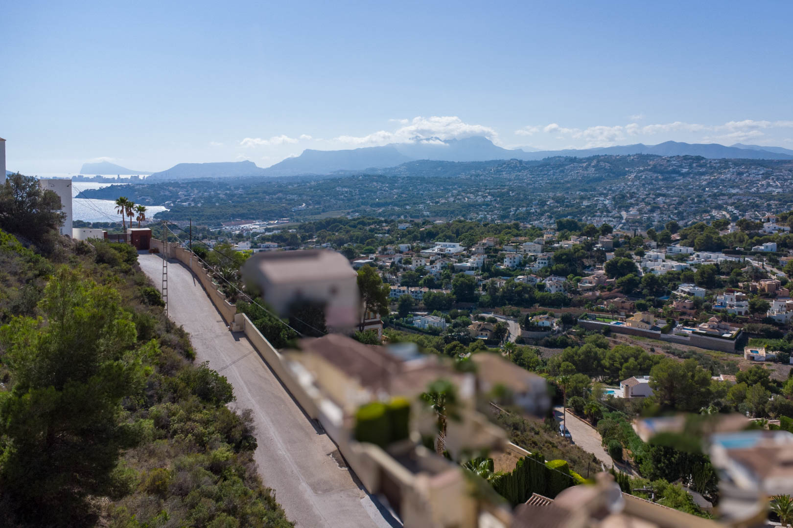 Panoramisch uitzicht vanaf het hooggelegen perceel in El Portet, met zicht op de woonwijk, de kustlijn van Moraira en de verre bergketens onder een heldere hemel.