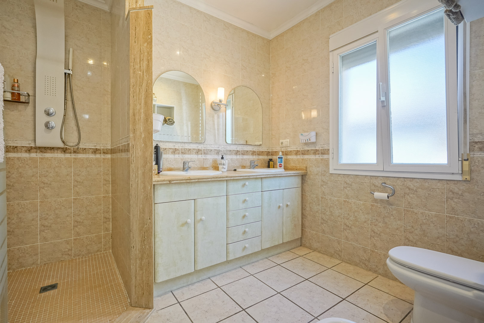 Badkamer met inloopdouche, wastafelmeubel met dubbele wasbak en crèmekleurige tegels.