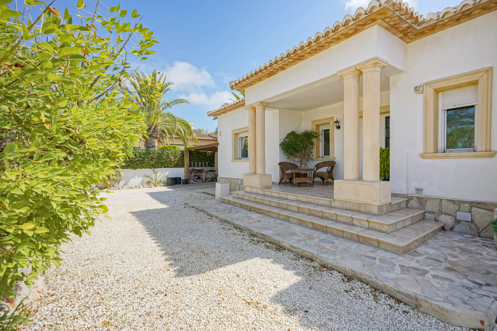 Villa-ingang met wit grind, veranda met kolommen en rotan meubilair.