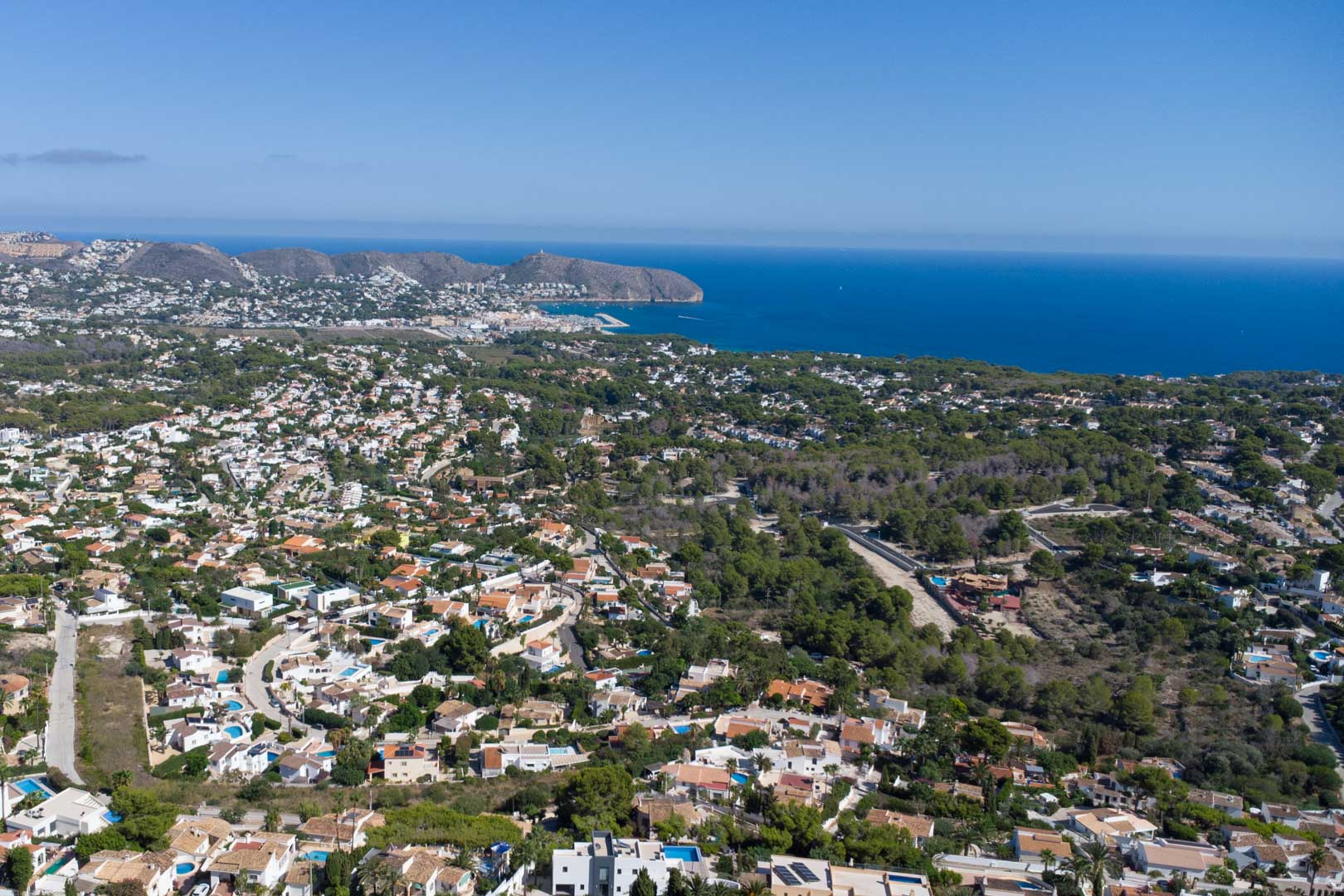 Panoramisch luchtfoto van de kustlijn van Moraira, met de jachthaven en Cap d'Or, omgeven door woonwijken en mediterrane dennenbossen.