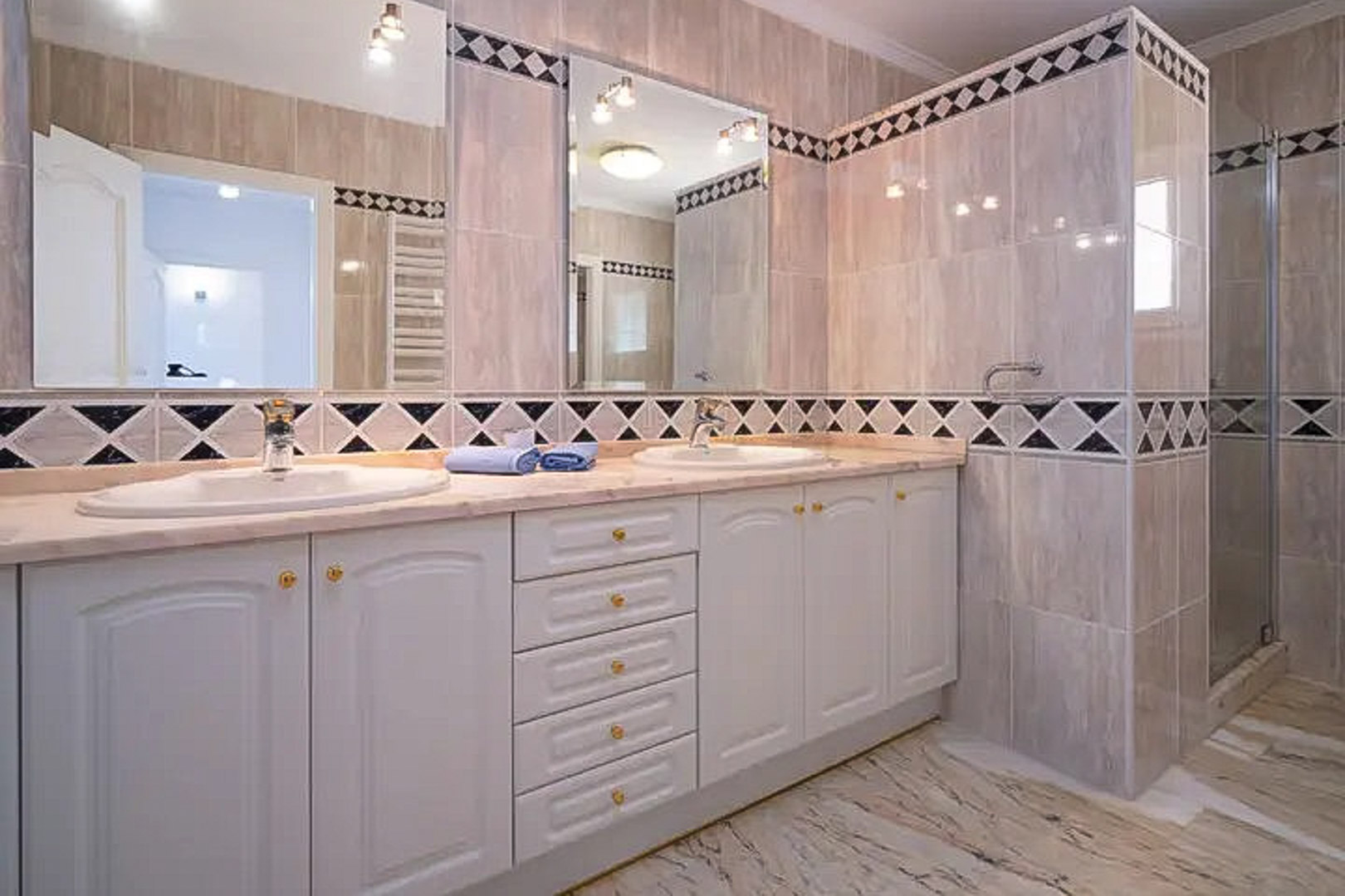 Badkamer met dubbele wastafel, wit klassiek meubel en marmeren blad. Voorzien van decoratieve ruitvormige tegels en een ruime inloopdouche aan de rechterzijde.