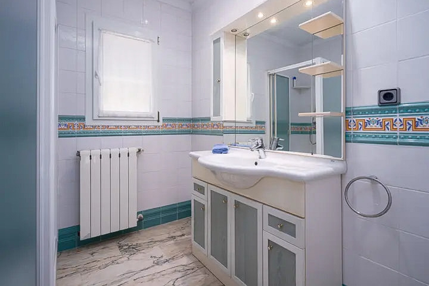 Badkamer met witte wandtegels en mediterrane sierrand, wastafelmeubel met matglazen deurtjes, grote verlichte spiegel en een marmeren vloer.