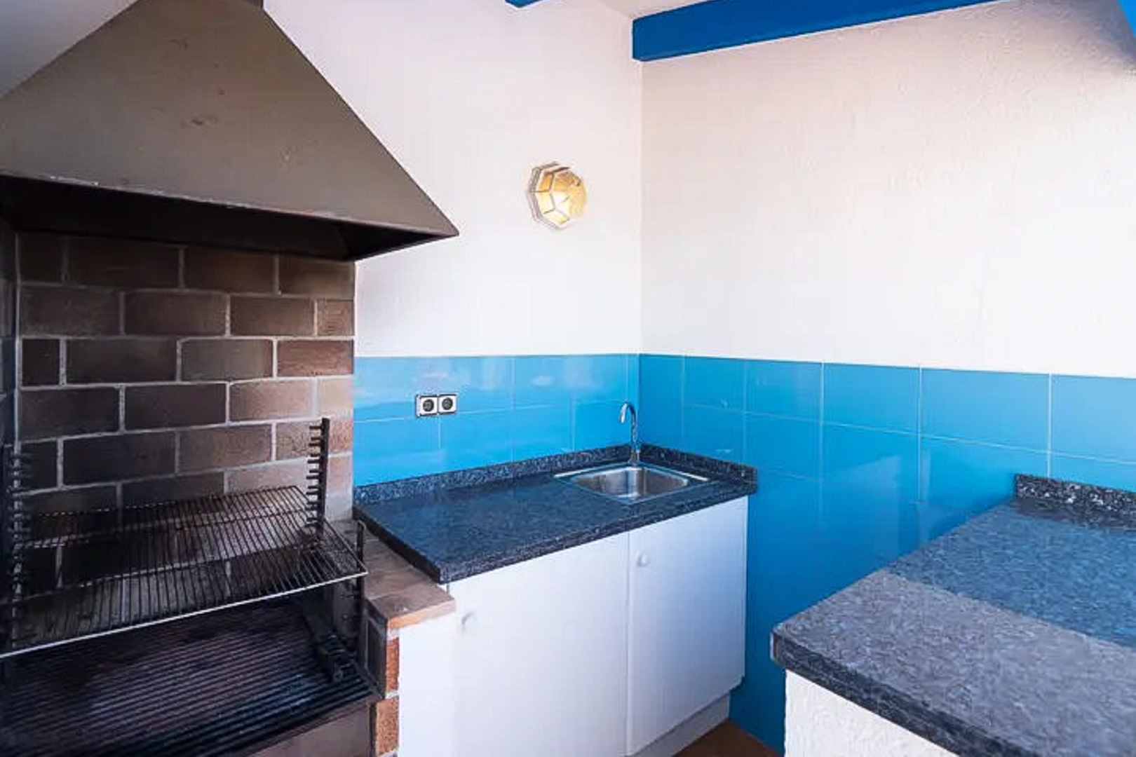 Buitenbarbecue met metalen afzuigkap, donker granieten werkblad, roestvrijstalen spoelbak en mediterrane blauwe tegels.