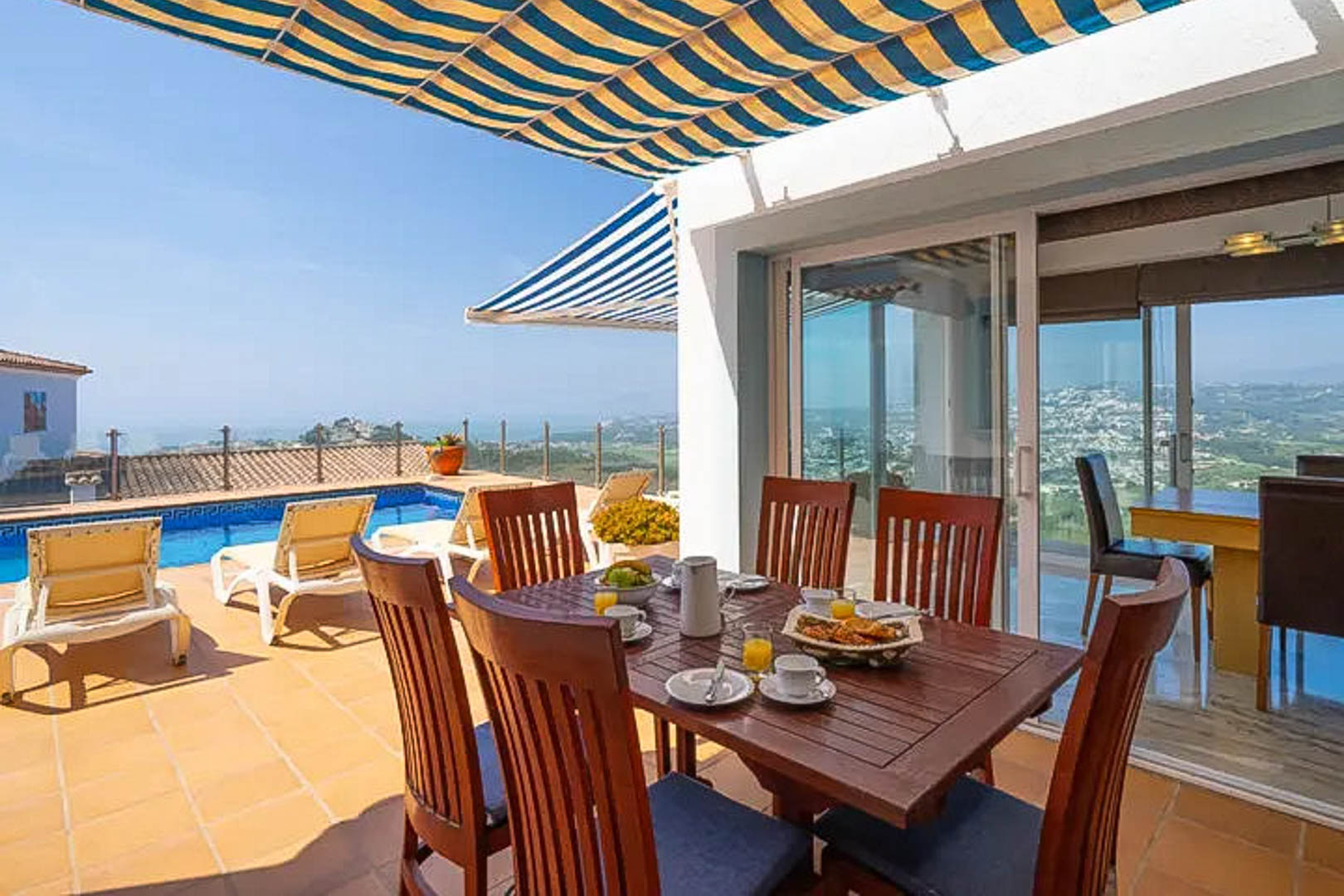 Buitenterras met houten eethoek onder een gestreept zonnescherm, uitkijkend op het privézwembad en de panoramische kustlijn van Moraira.